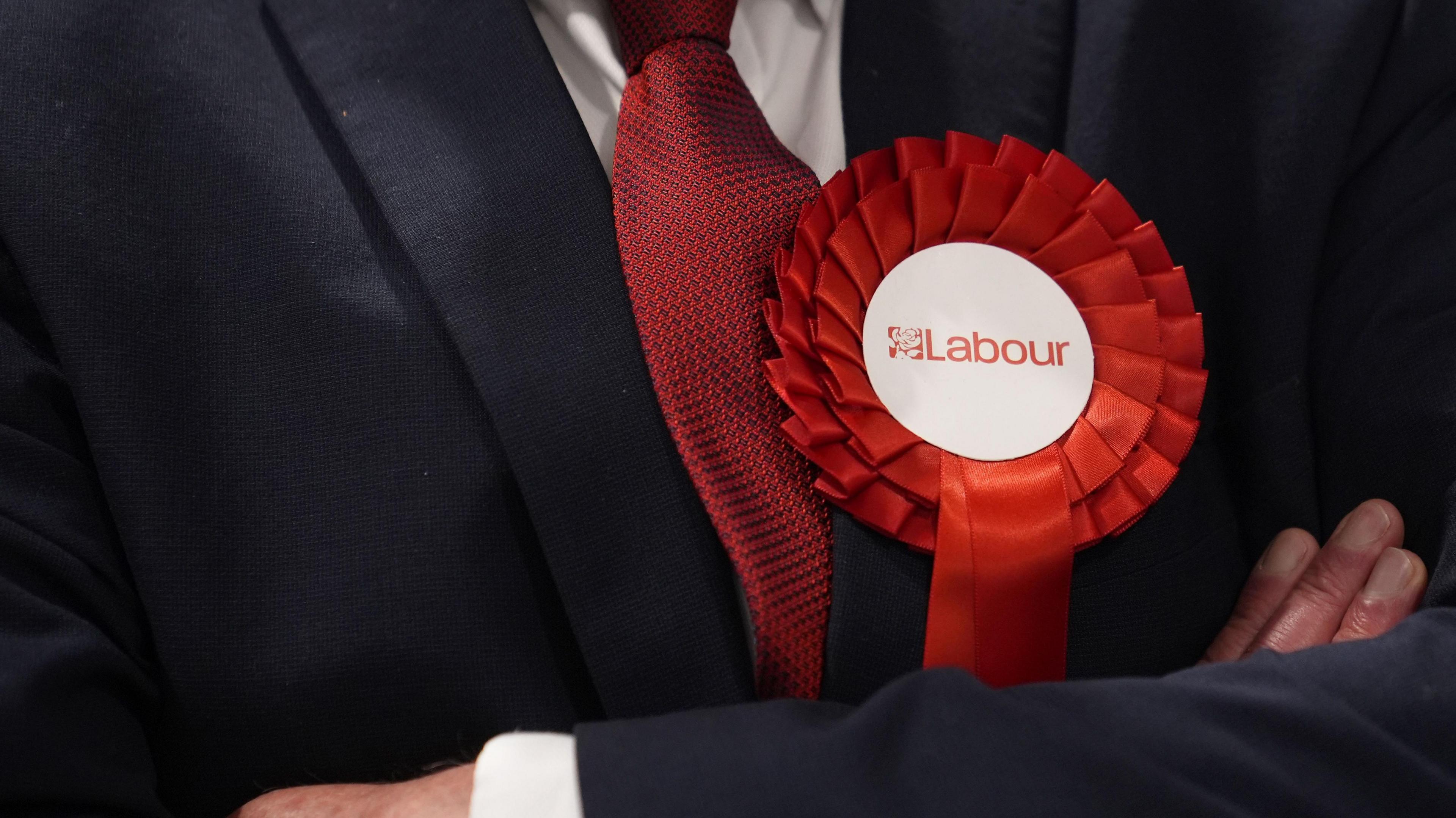 a labour rosette on a person wearing a black suit and red tie with their arms crossed