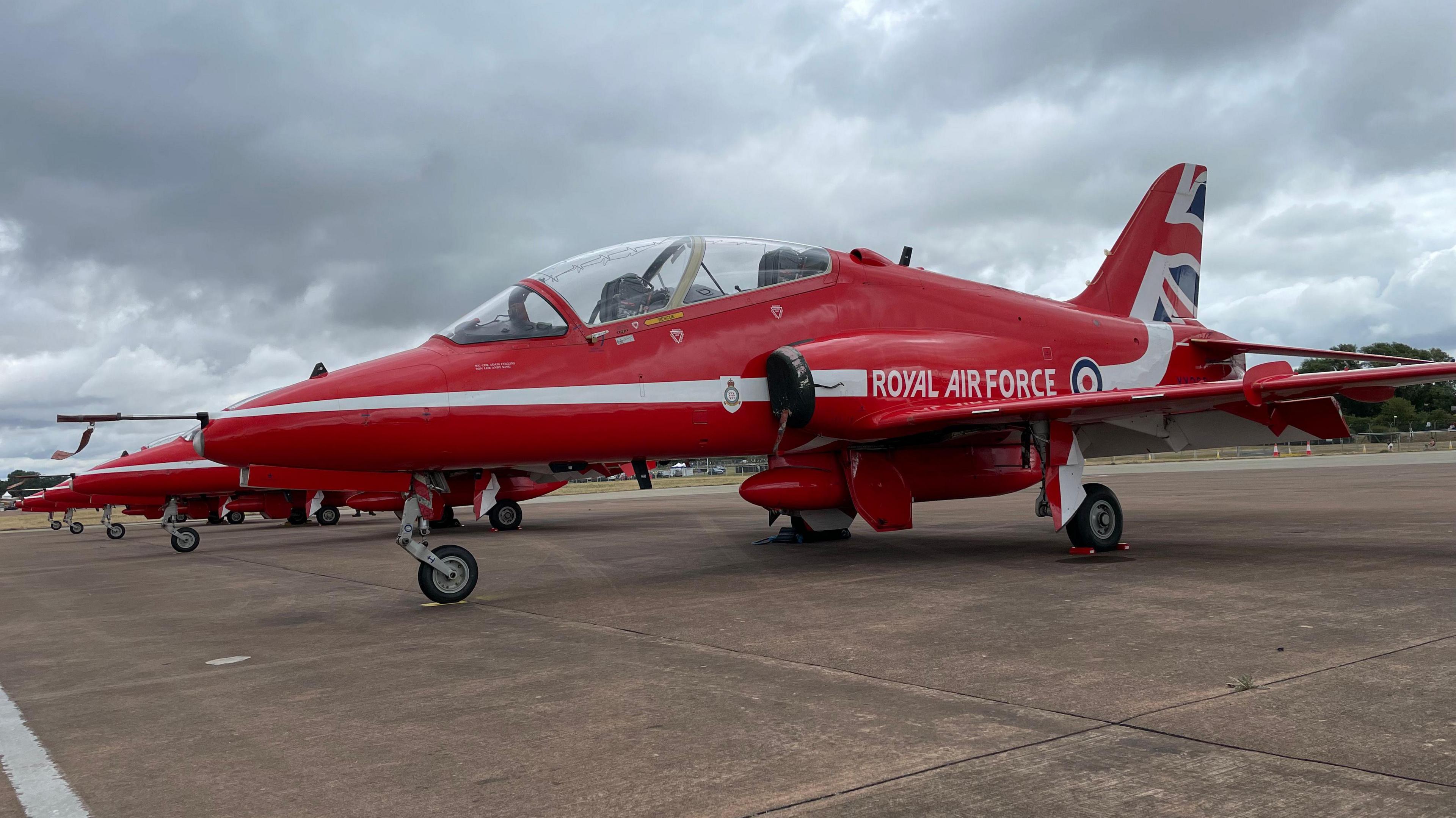 A close-up of a bright red Red Arrows jet on the ground with the Royal Air Force name on the side and a number of other jets lined up beside it