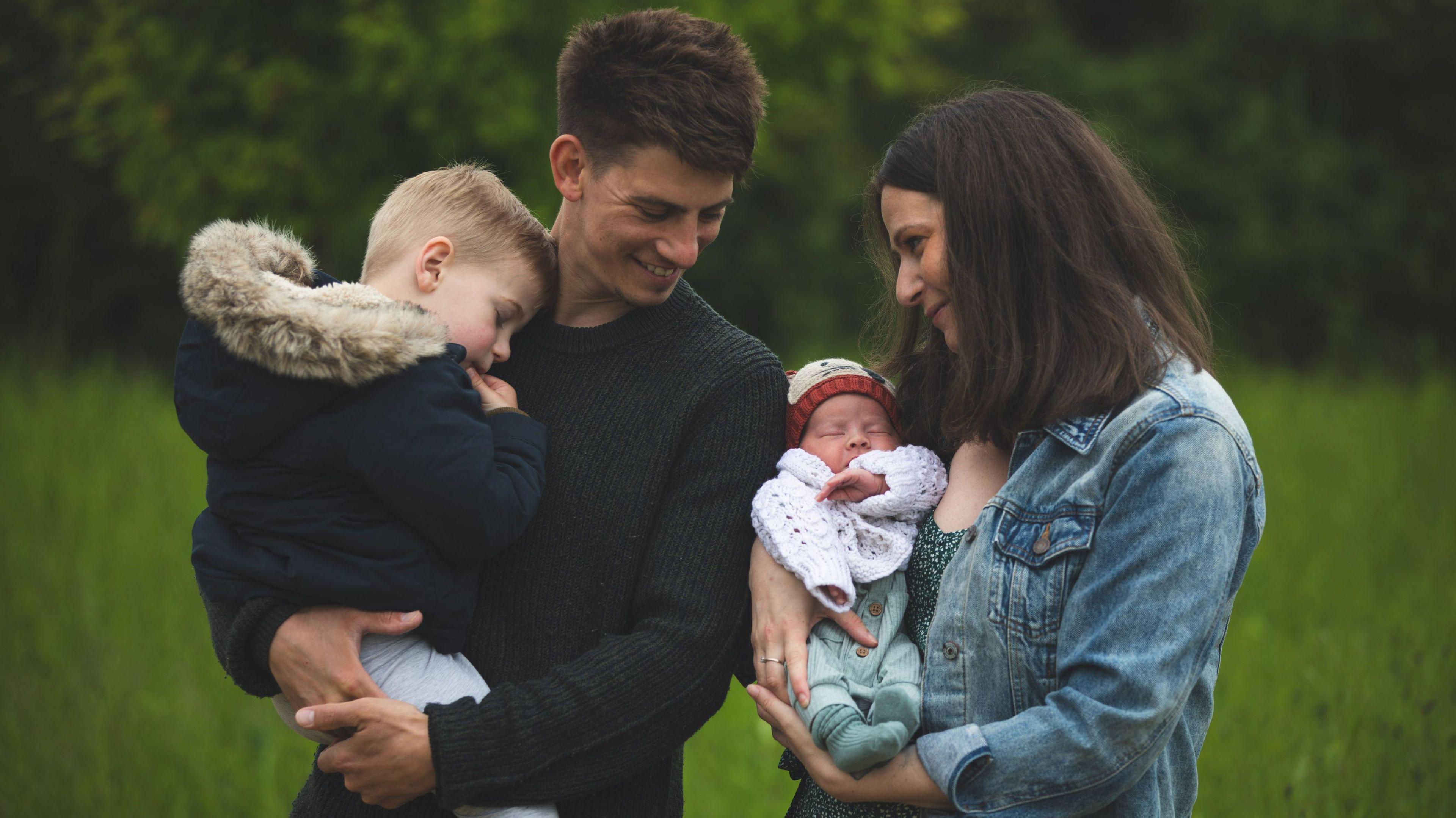 Sophie and her husband holding their two boys in their arms in a field smiling at each other