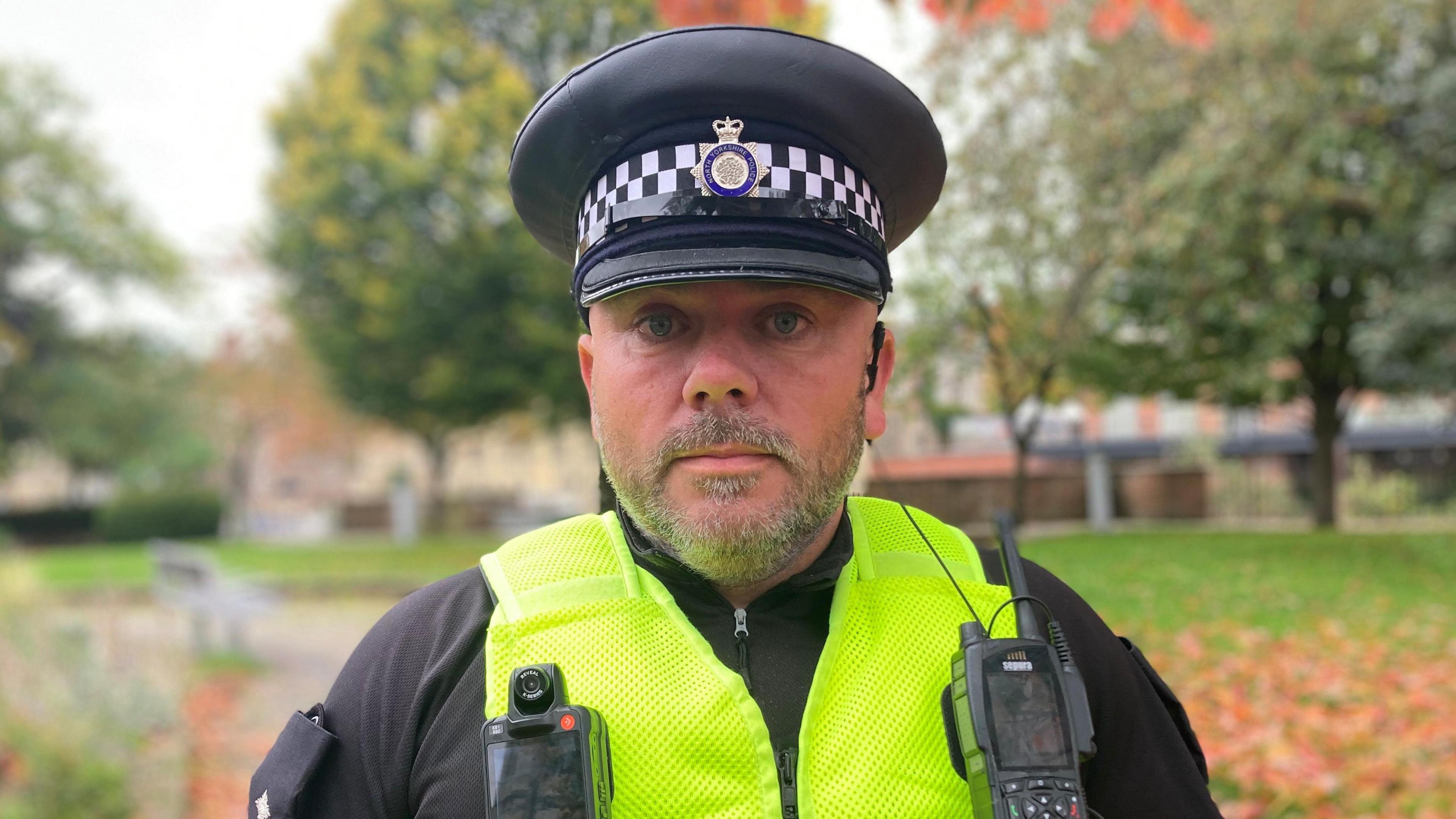 Insp Nick Plumb, from North Yorkshire Police, is wearing a police issue cap, black clothing with a yellow police vest with a body camera and police radio attached to it