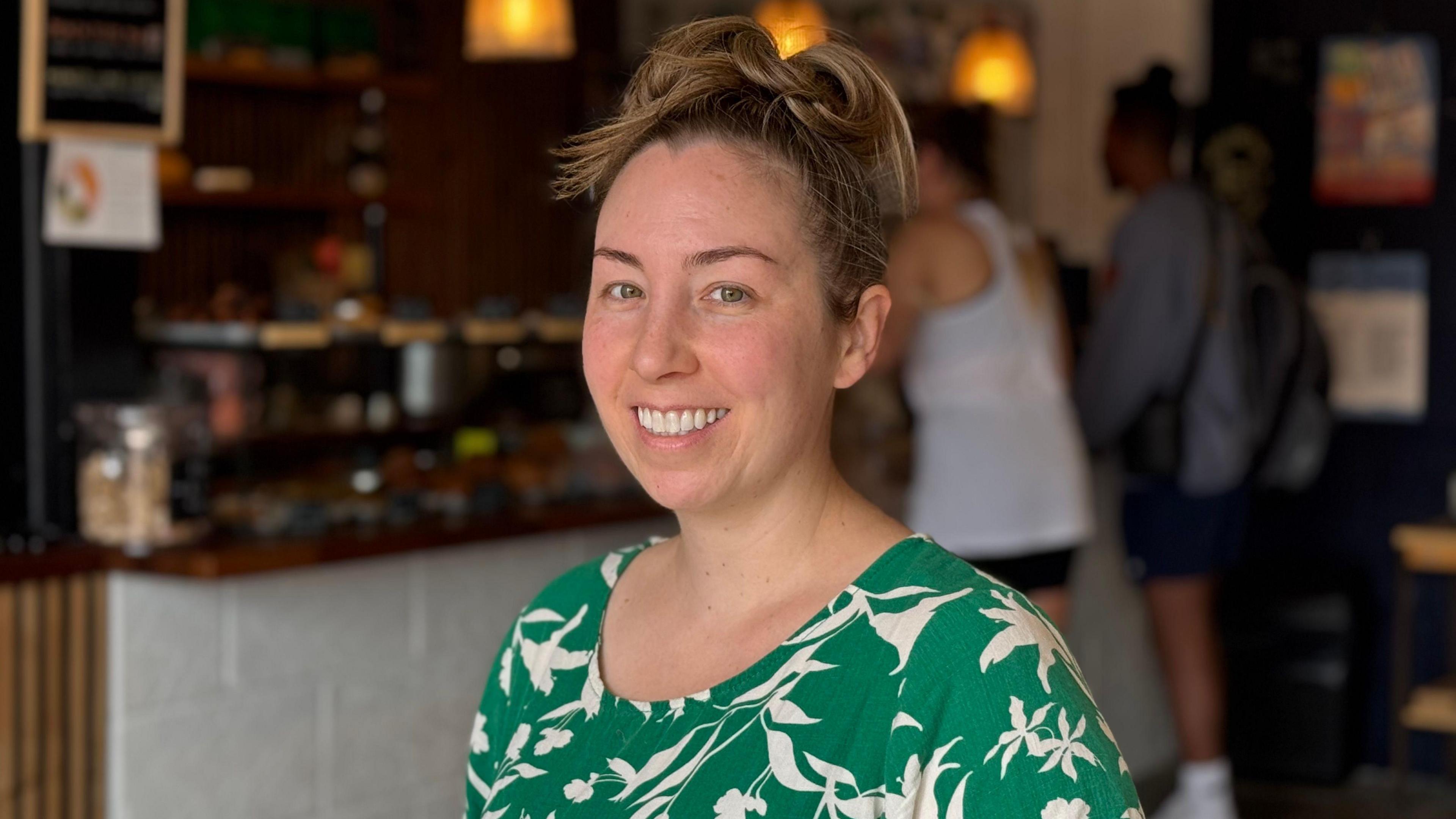 Heather is smiling at the camera. She has her hair tied up and is wearing a green and white dress. She is in the cafe in Northstowe. There are out of focus customers in the background, buying coffee from the barrista.
