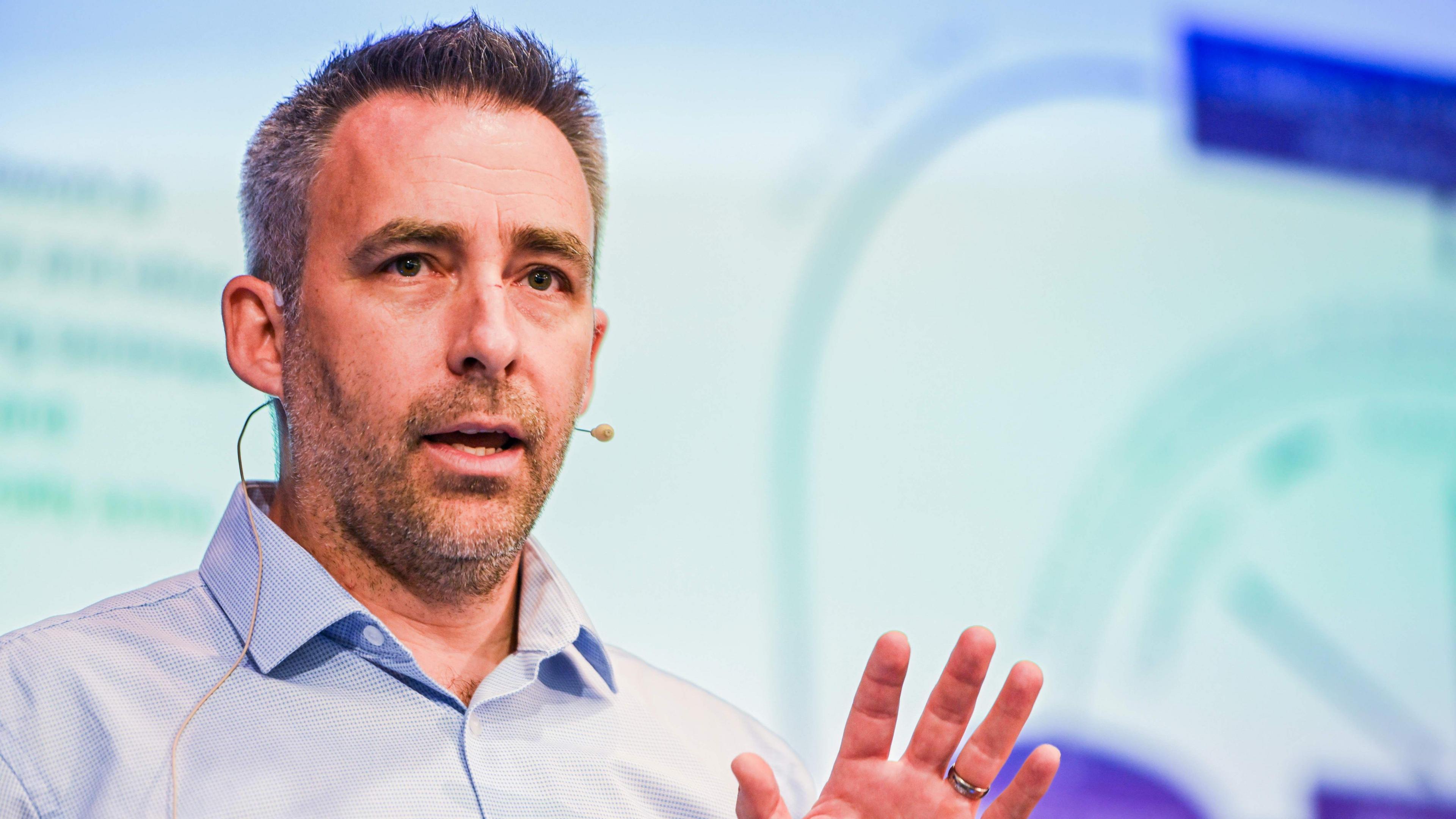 A man in a light blue shirt talking into a small microphone attached to his ear
