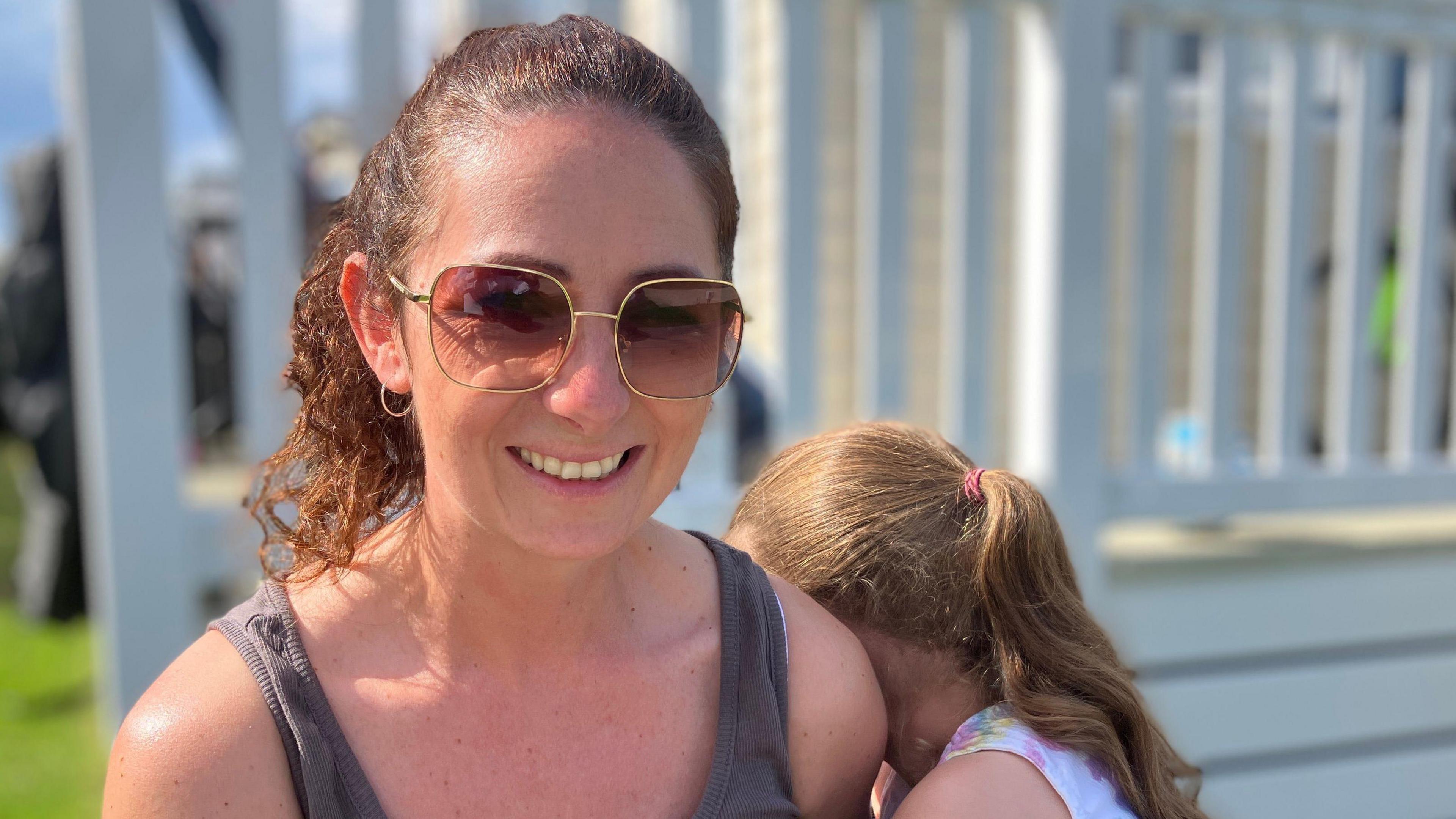Natalie Bass, dynes gyda gwallt brown wedi ei dynnu'n dynn am ei phen, yn gwenu tua'r camera. Mae ei merch fach wrth ei hochr, yn cuddio'u hwyneb yn ei hysgwydd ac mae'r ddwy'n eistedd o flaen grisiau gwyn eu carafán