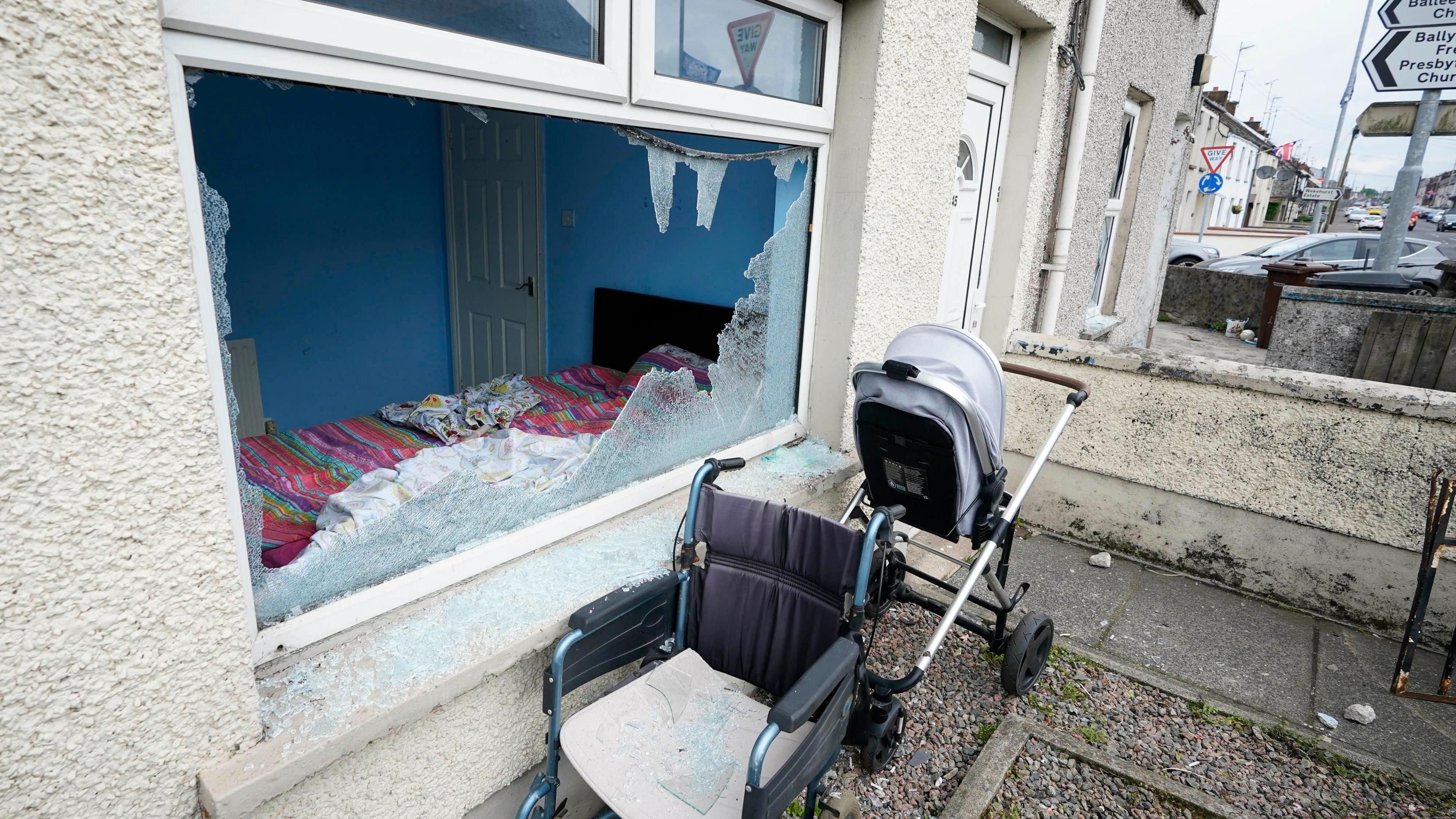 A black wheelchair and white pram sit outside a smashed ground floor window. On the other side of the smashed glass is a blue painted bedroom, with a black bedframe and pink bed sheets.