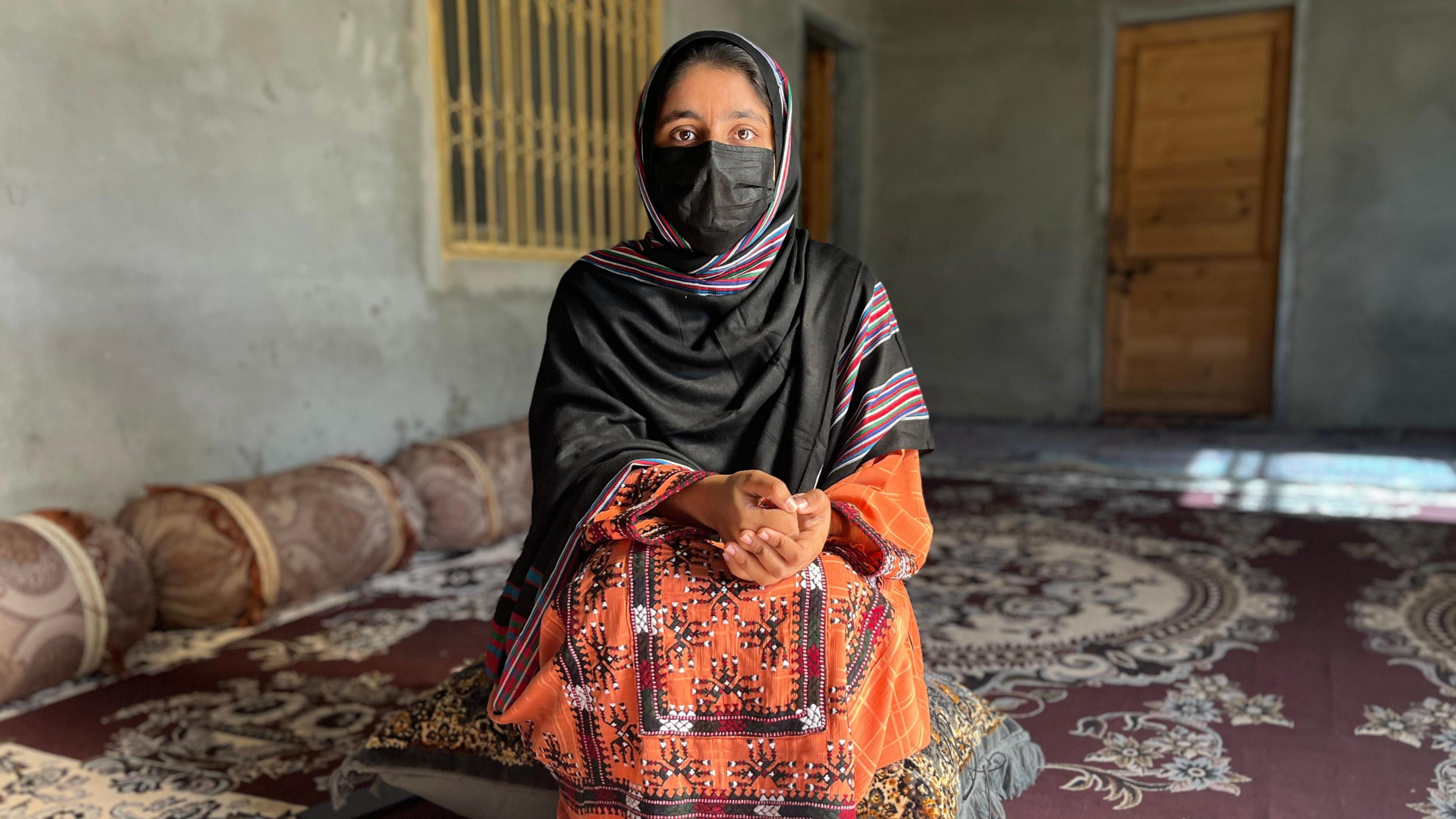 A young woman with her face covered looks at the camera, while sitting on a grey floor cushion 