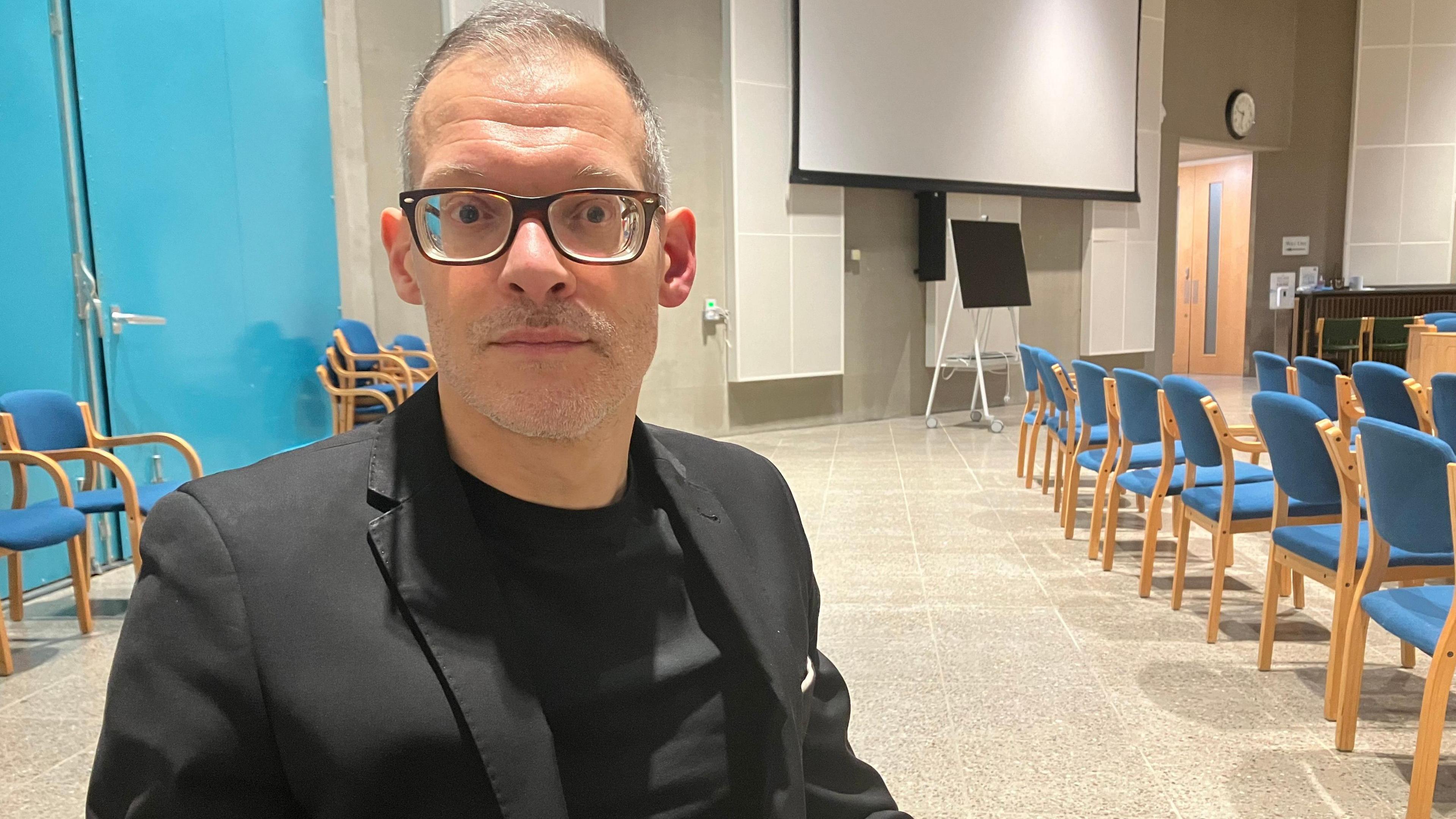 Independent Central Bedfordshire councillor Adam Zerny, who has glasses and is wearing a black suit jacket and a black t-shirt. He has short grey hair. He is sitting down. There are rows of blue and brown chairs in the background.