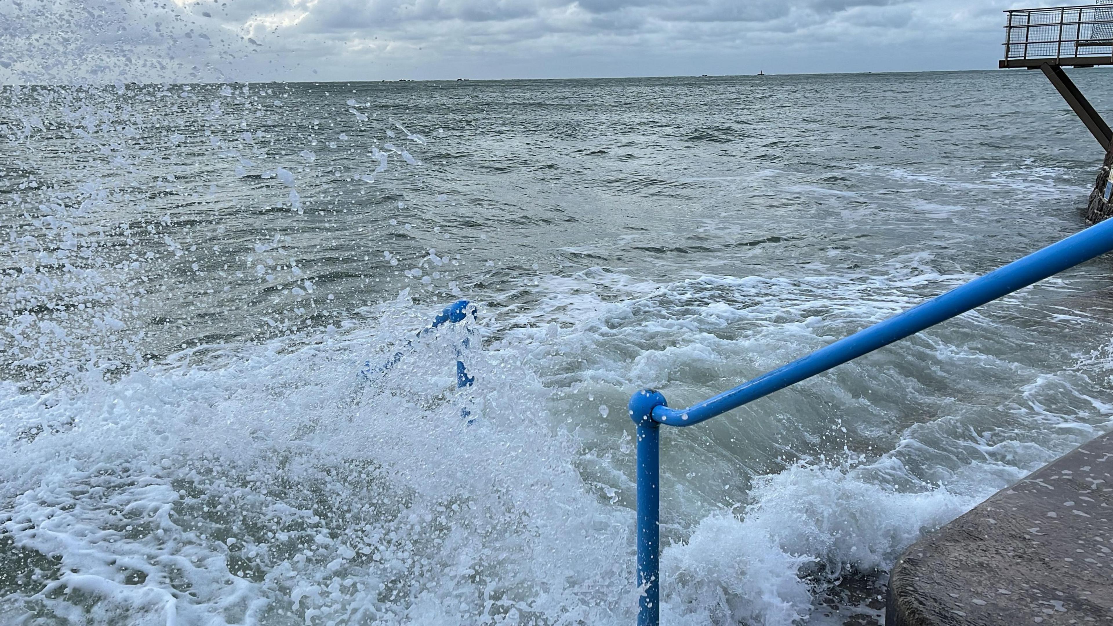 A wave crashes along railings and steps at the Lido in Harve des Pas along the seafront in Jersey during stormy weather. The railings leading into the water are blue.