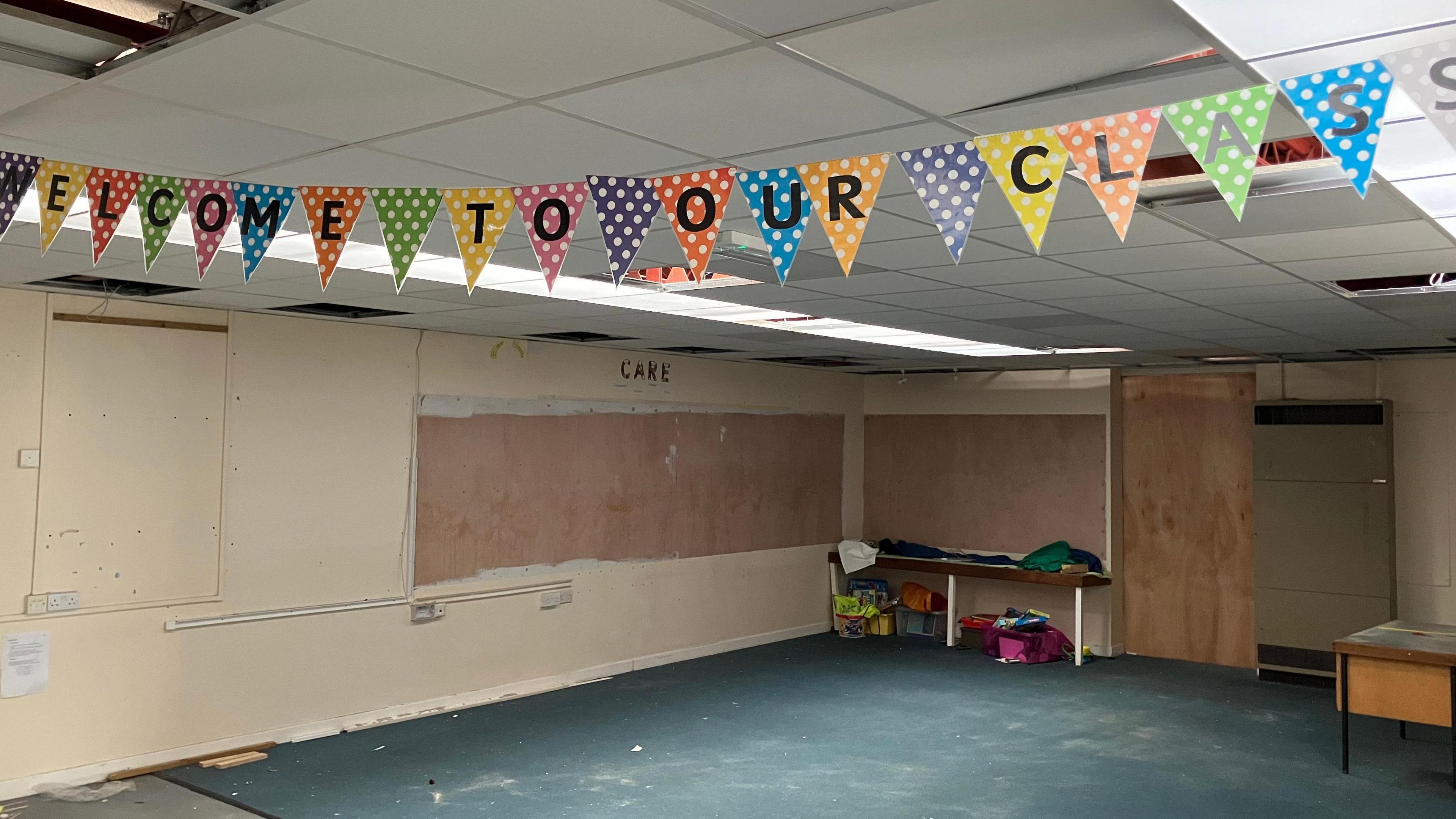 A general view of a derelict classroom. There are no desks or chairs or carpet. A banner that reads "Welcome to Our Class" is still attached to the ceiling.