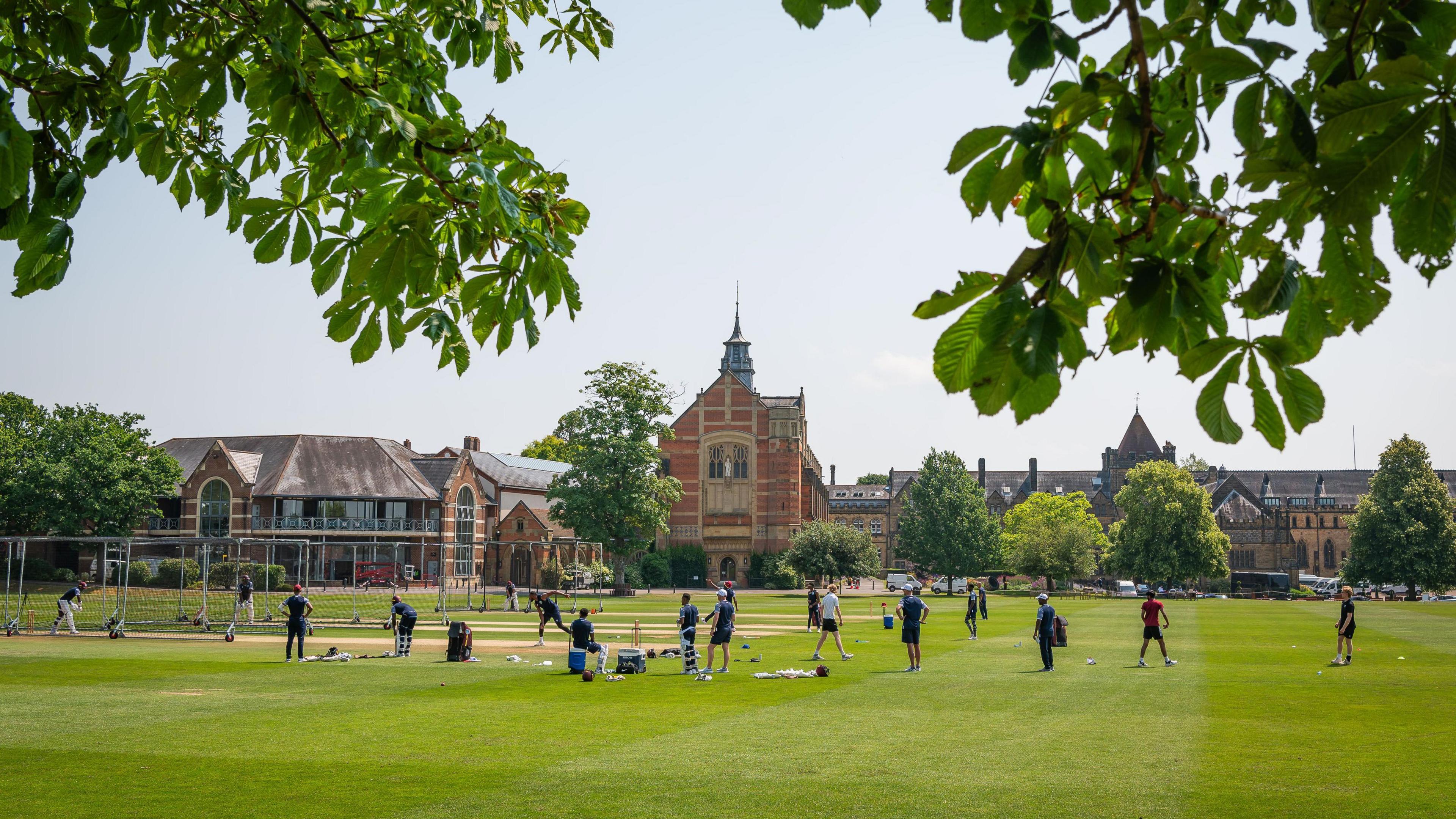 A group of people stood on a grass field. In the background there is a brown brick building.