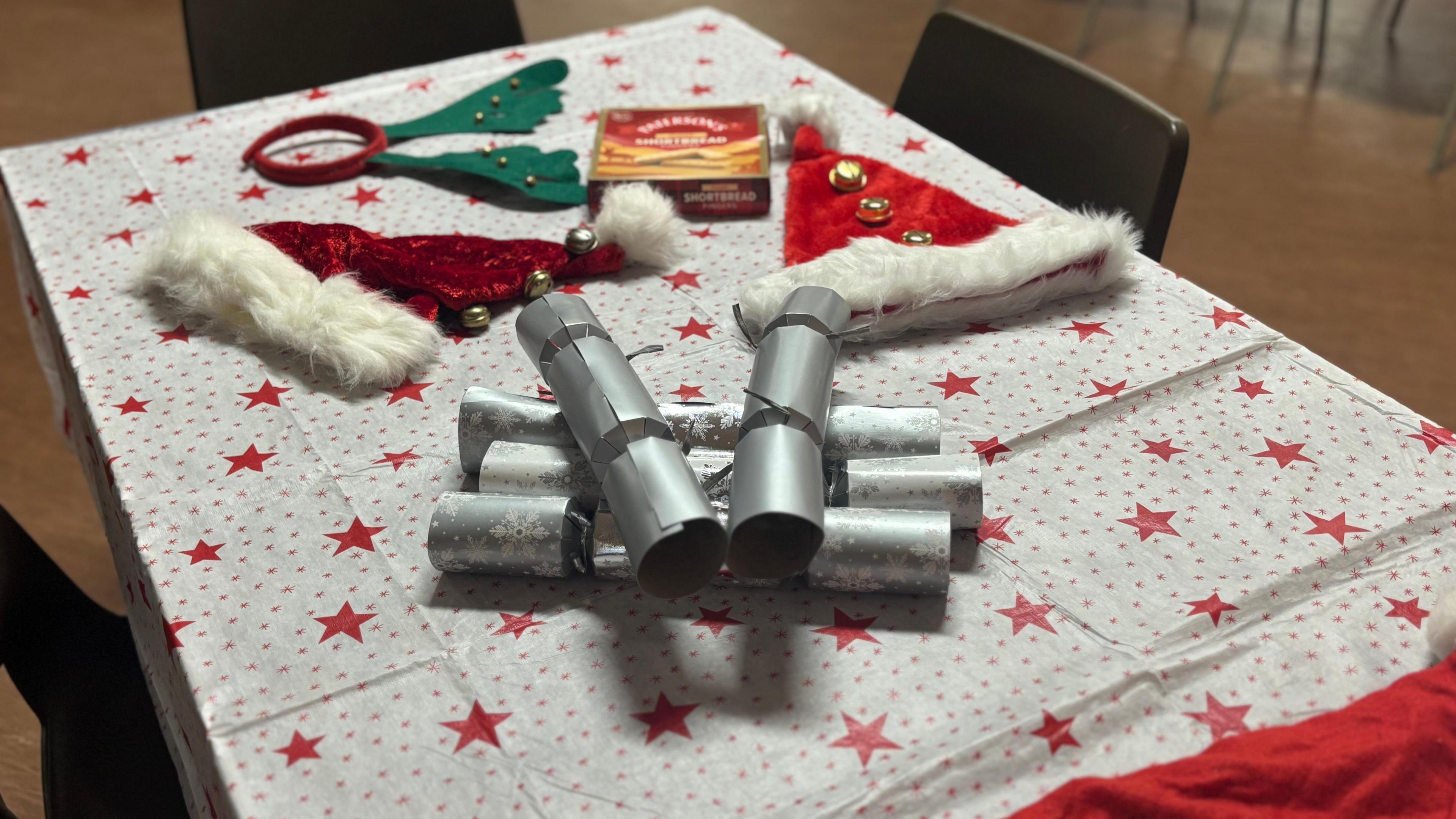 On a table are Christmas crackers, two santa hats, a box of shortbread and some reindeer antlers on a white and red tablecloth with stars on it. Around the table are plastic brown chairs.