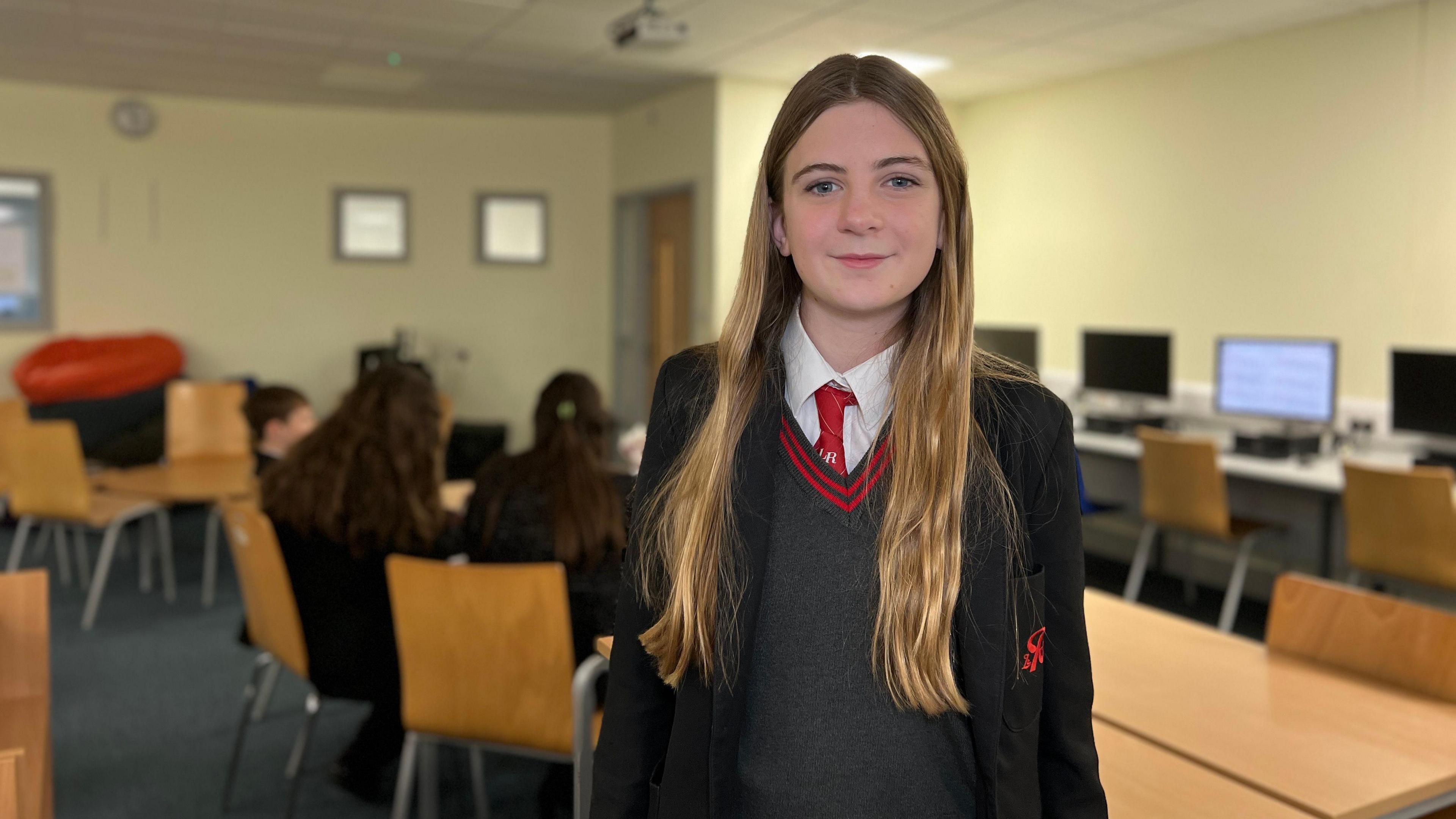 Georgia looks at the camera and she has long light brown hair which comes down to her chest. She has a black jacket on with the school logo on it and a grey jumper underneath with a red tie too.