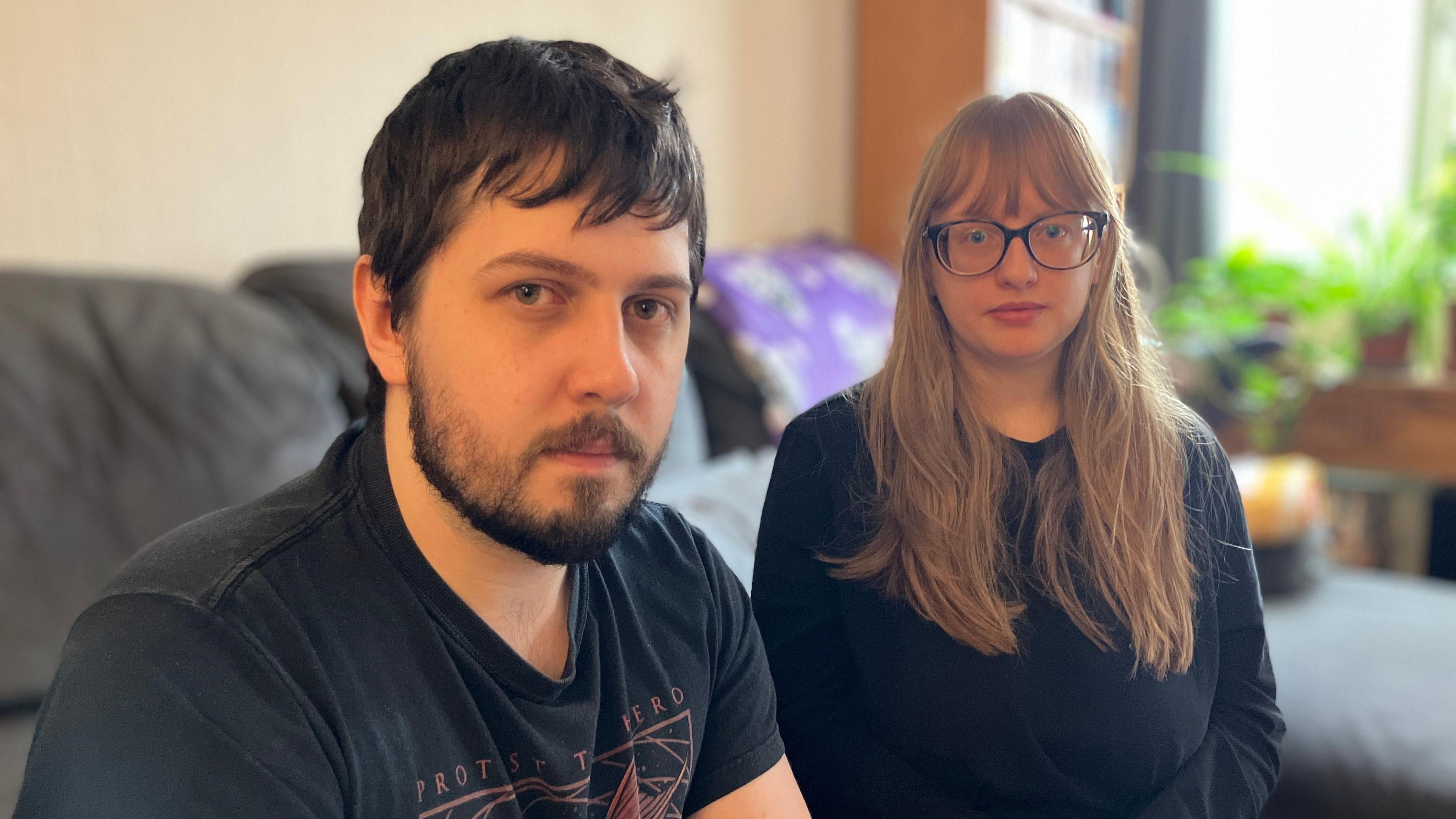 A man with dark hair and a beard wearing a T-shirt sitting on a sofa next to a woman with long hair and glasses.