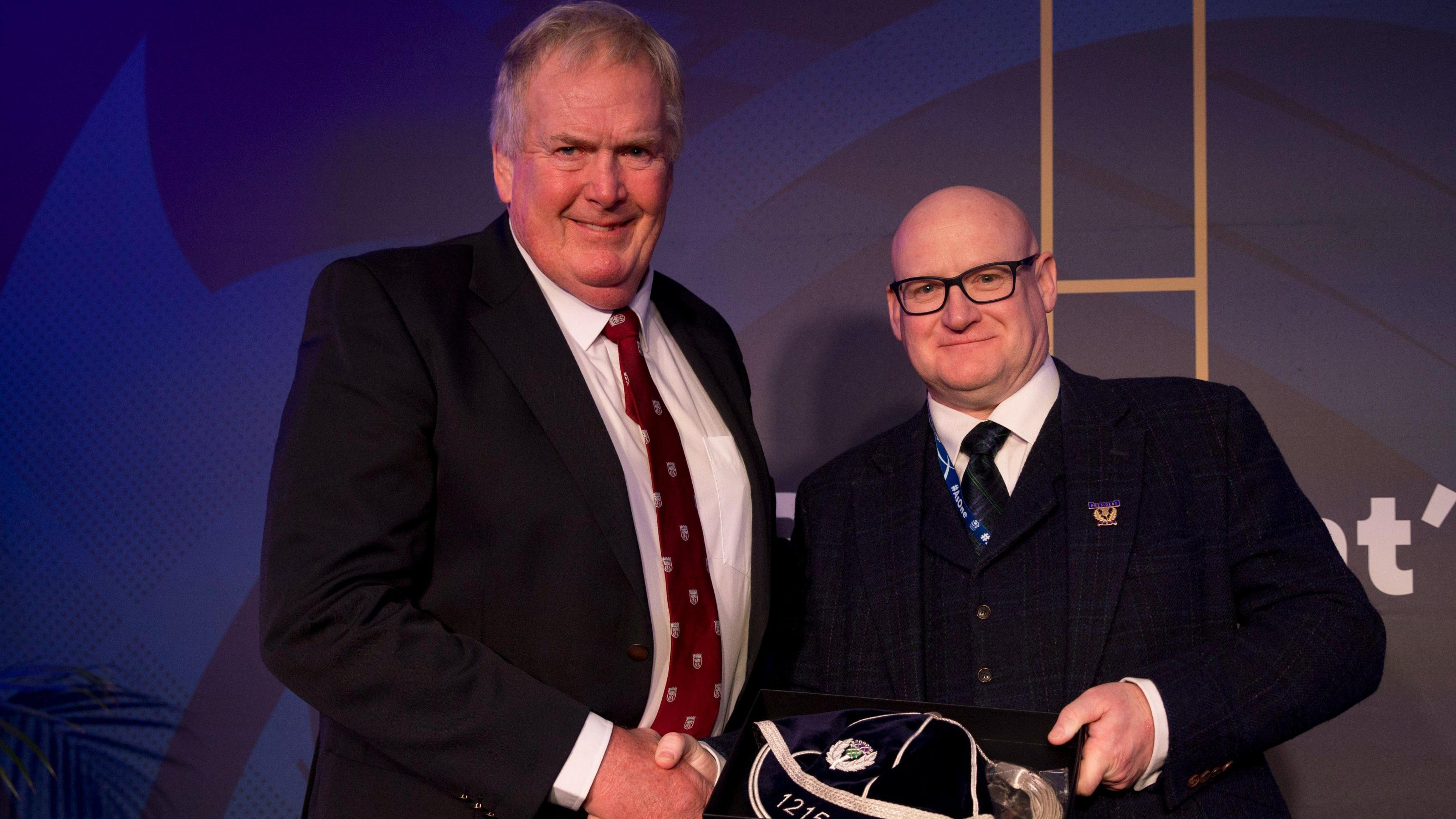 Two men in formal attire are standing and shaking hands. The man on the left has light brown hair and is wearing a dark navy suit jacket, a white shirt, and a maroon patterned tie. The man on the right is bald, has glasses, and is wearing a navy blue suit jacket.