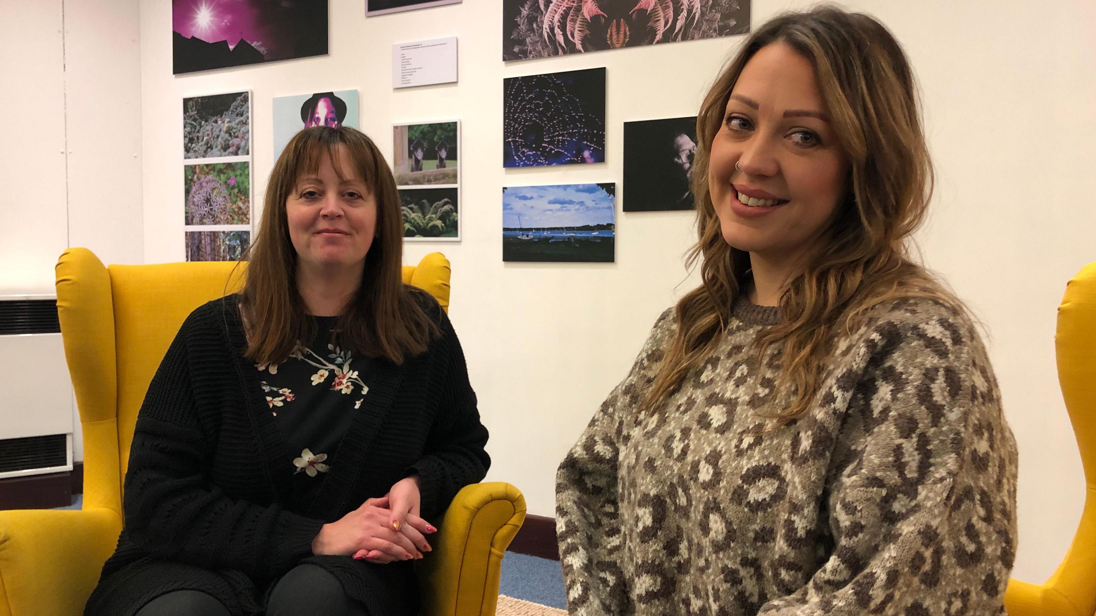 Michelle and Charlotte sit on two yellow arm chairs in front of photography exhibition inside a library