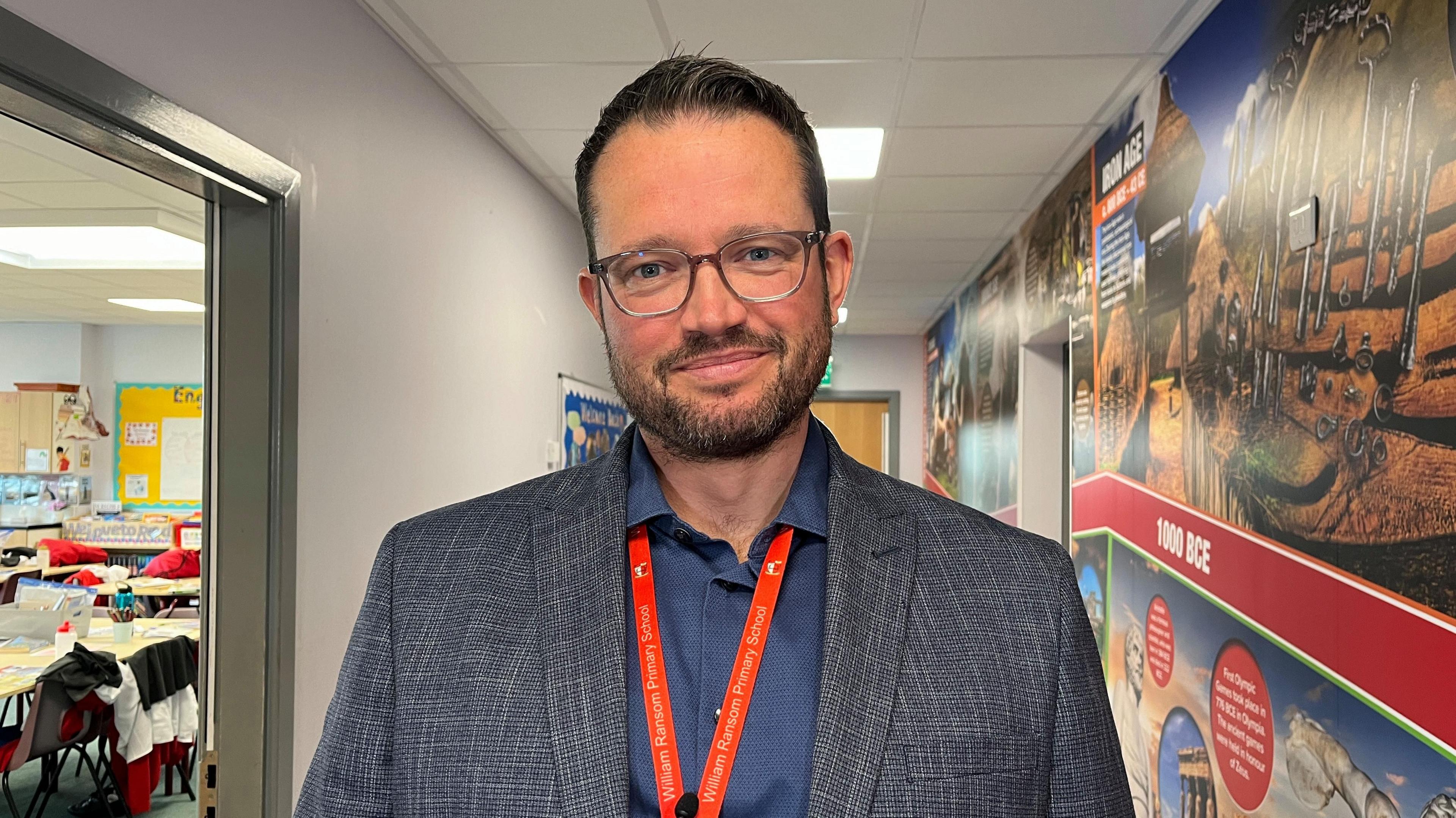 A man smiling in a school corridor. He is wearing glasses, a grey blazer and a navy shirt. He has a red lanyard with William Ransom Primary School written on it.