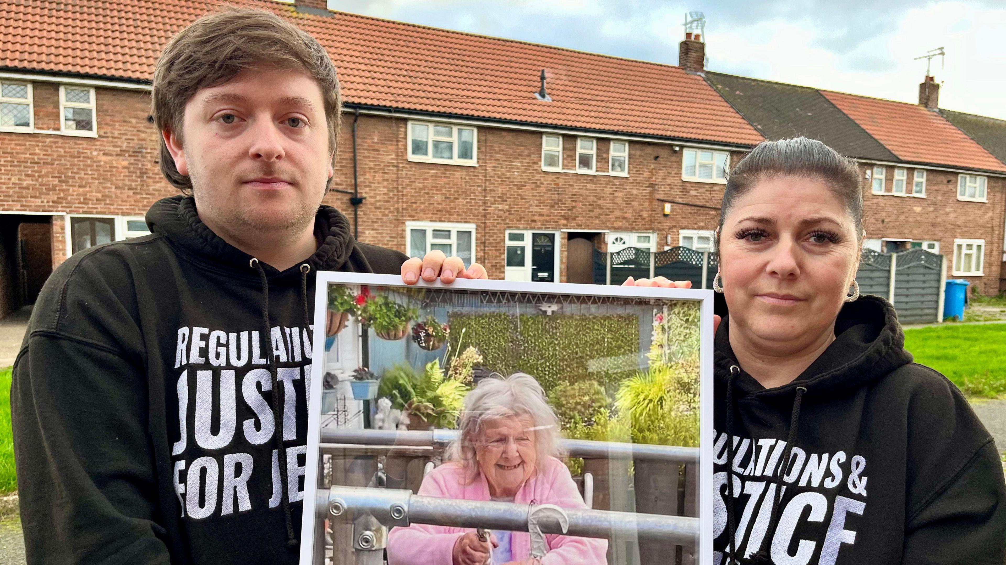 Tristan Essex and Claire Stockdale are standing outside a row of terraced houses and holding a large framed photo of their grandmother. They are wearing black hoodies printed with white text, which reads: "Regulation and justice for Jessie".