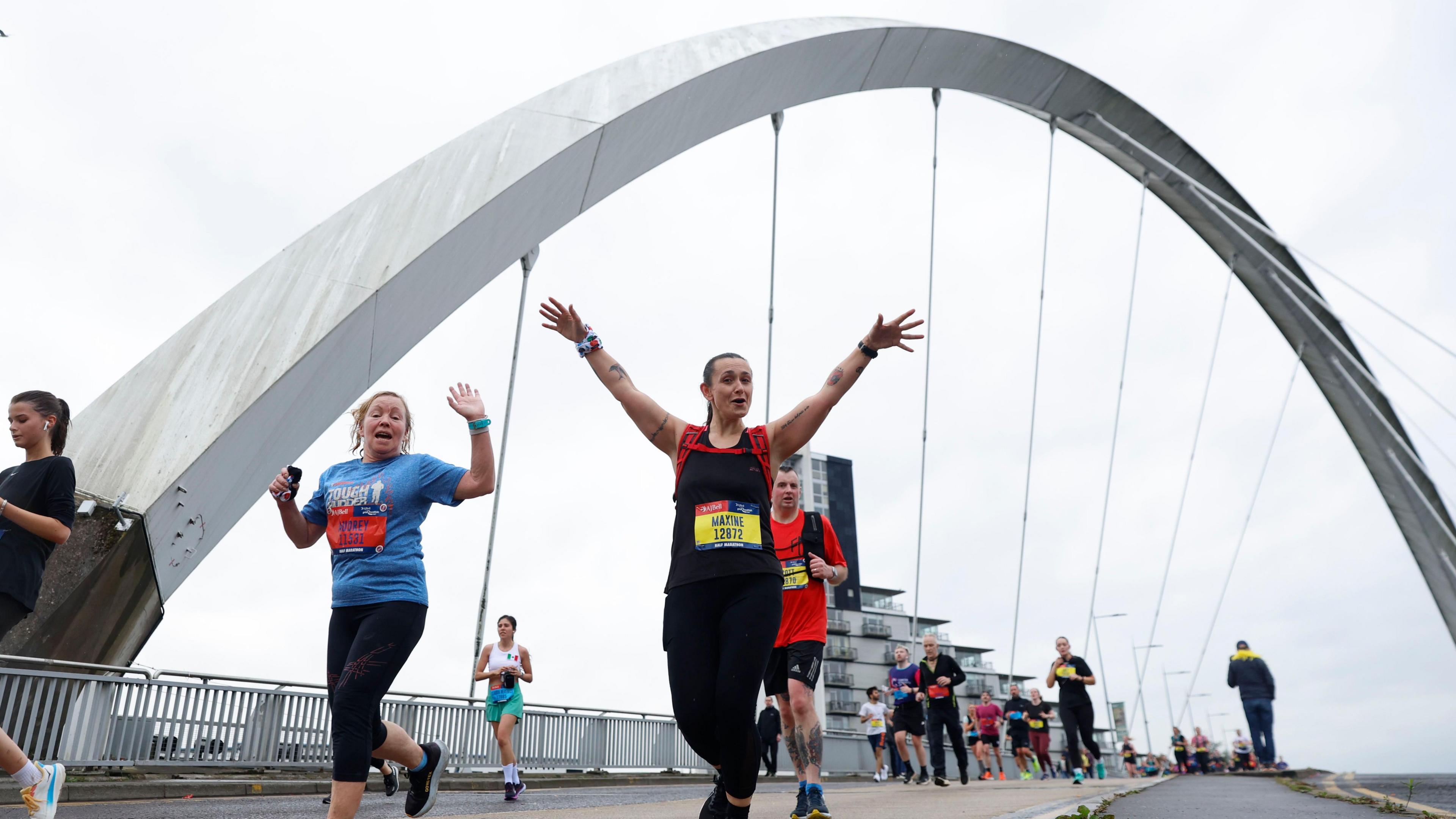 Runners going over the squinty bridge in Glasgow. Two women are the focus of the photo. One on the left is wearing a blue top and waving at the camera. The lady on the right is waving her hands in the air.