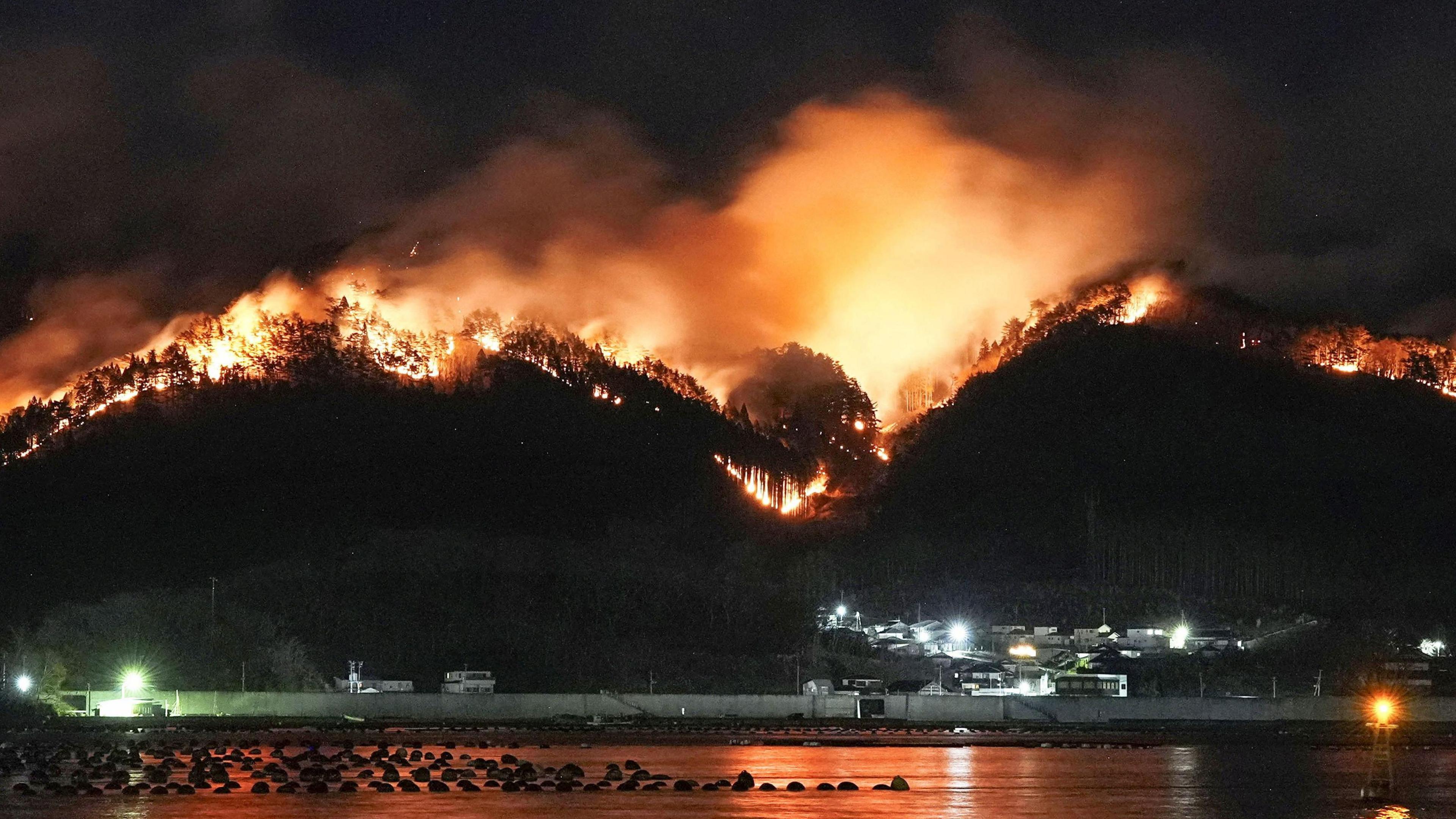 In the foreground water can be seen in a harbour, while the hills behind are on fire. 