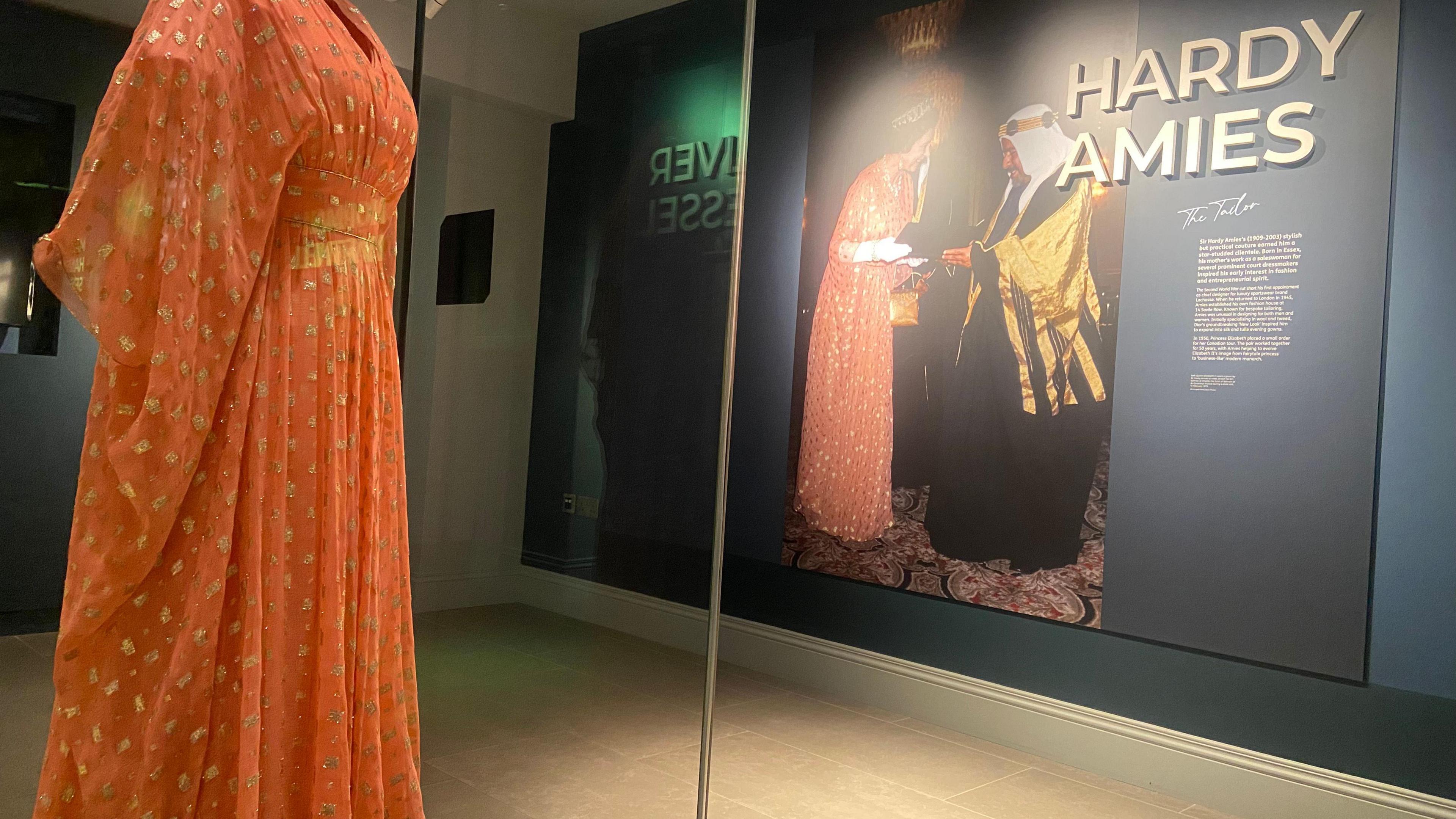 An evening dress worn by Queen Elizabeth II in 1979. It is a salmon-pink silk chiffon dress with small gold rectangles and bat-wing sleeves, encased in a glass cabinet. An exhibition sign can be seen in the background with a picture of Queen Elizabeth II wearing the dress at a state visit to Bahrain in 1979 where she is meeting the Emir of Bahrain.