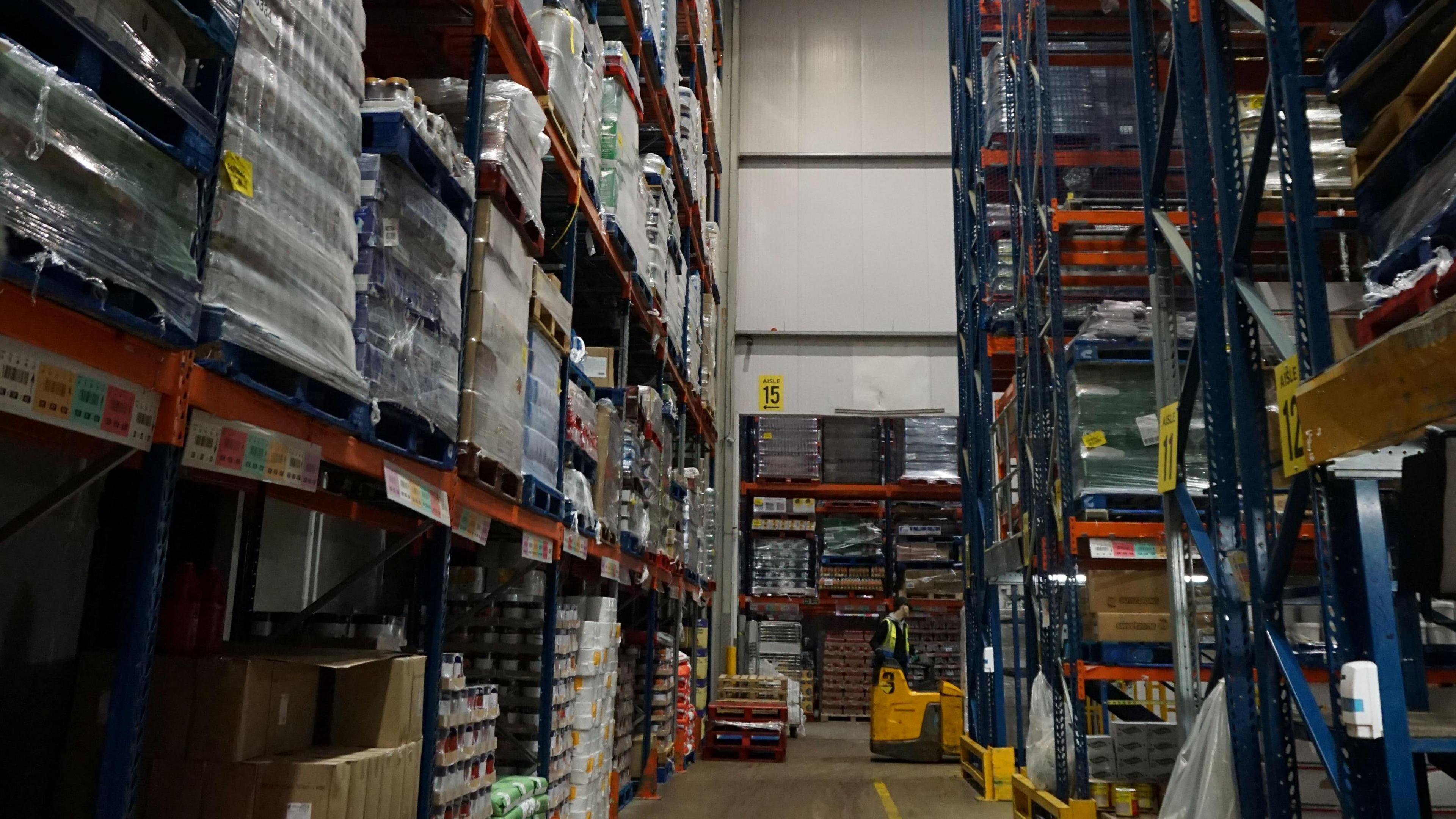 A warehouse with shelves of stock. There is a concrete floor with yellow markings.