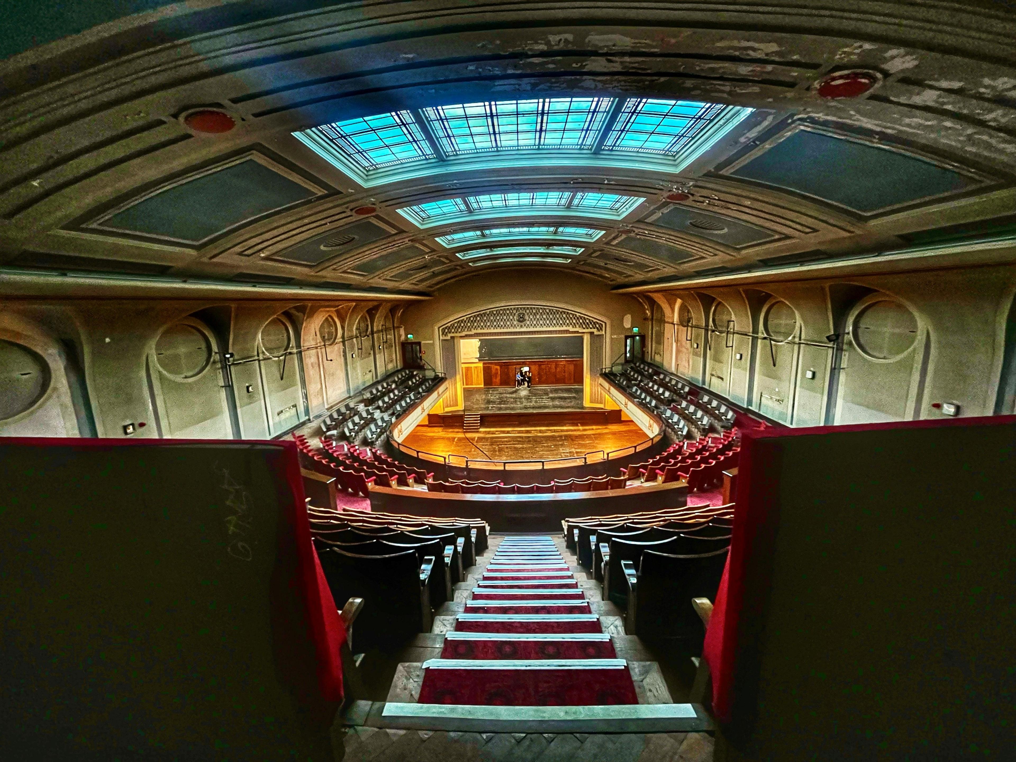 Old theatre photographed from opposite the stage. up the stairs at the very back. The theatre is empty and has red velvet seats, a slightly curved ceiling with skylight and light green walls.