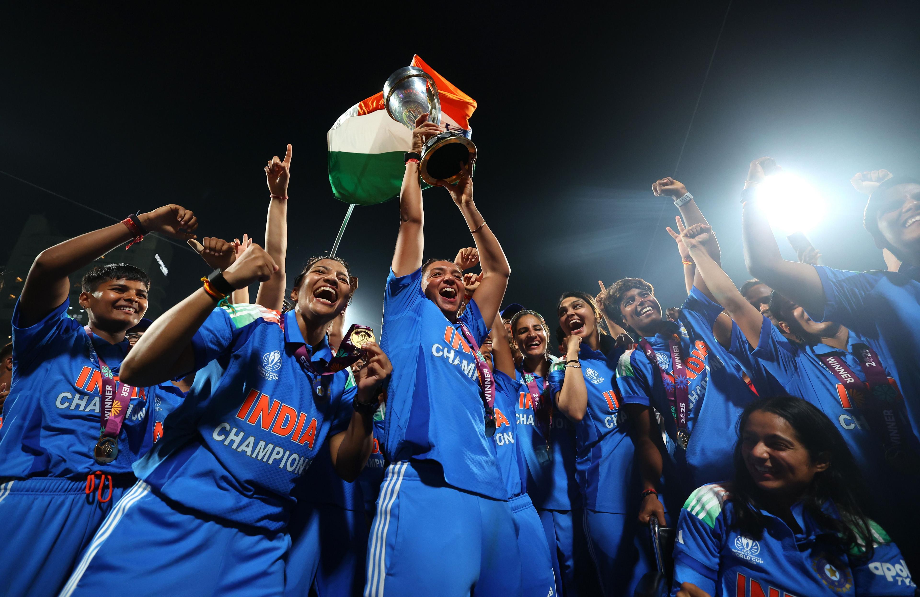 Indian Cricketers celebrate a win, wearing blue uniforms labeled “INDIA” and CHAMPIONS. One player lifts a trophy while others smile and display medals. The Indian flag and bright stadium lights highlight the moment.