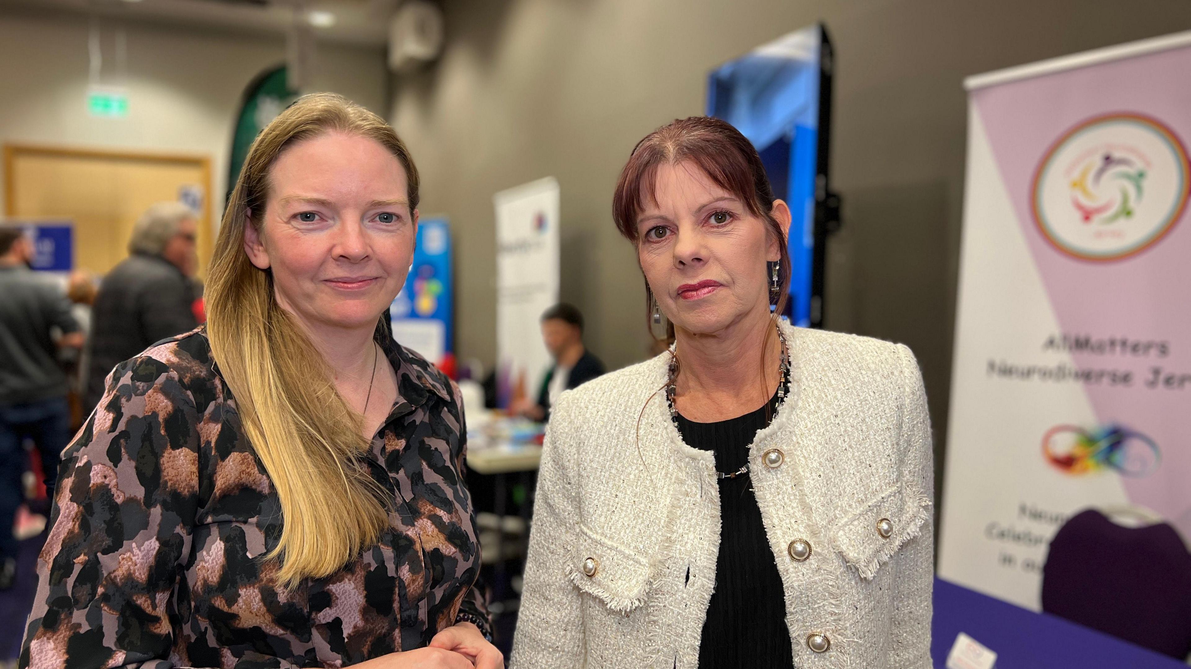 Niamh is wearing a black and patterned shirt and has long blond hair which comes down to her chest. Penny has a white jacket over a black top on and has dark red hair which is tied into a ponytail. Behind them, people are going to stalls being run by various charities.