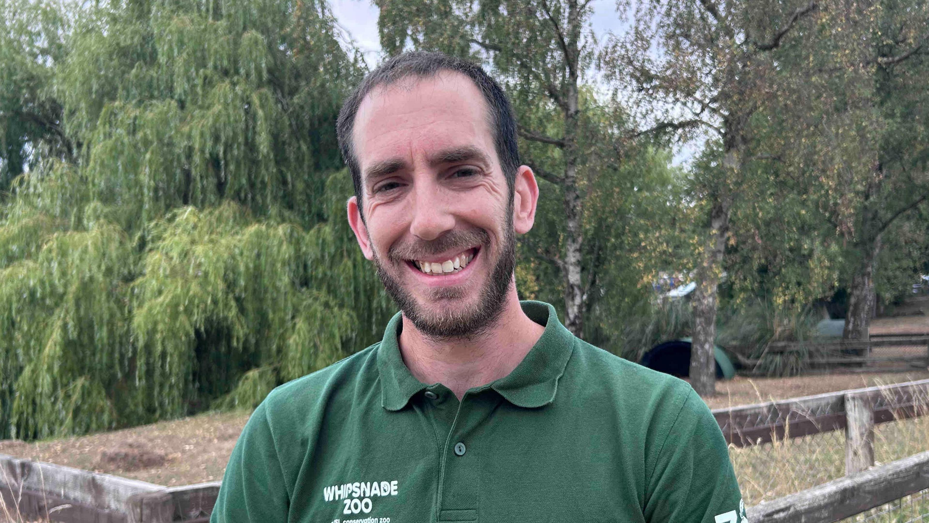 Kim Burton with short dark hair and beard, smiling at the camera and wearing a dark green Whipsnade Zoo T-shirt. There is grass and a wooden fence behind him and trees in the background