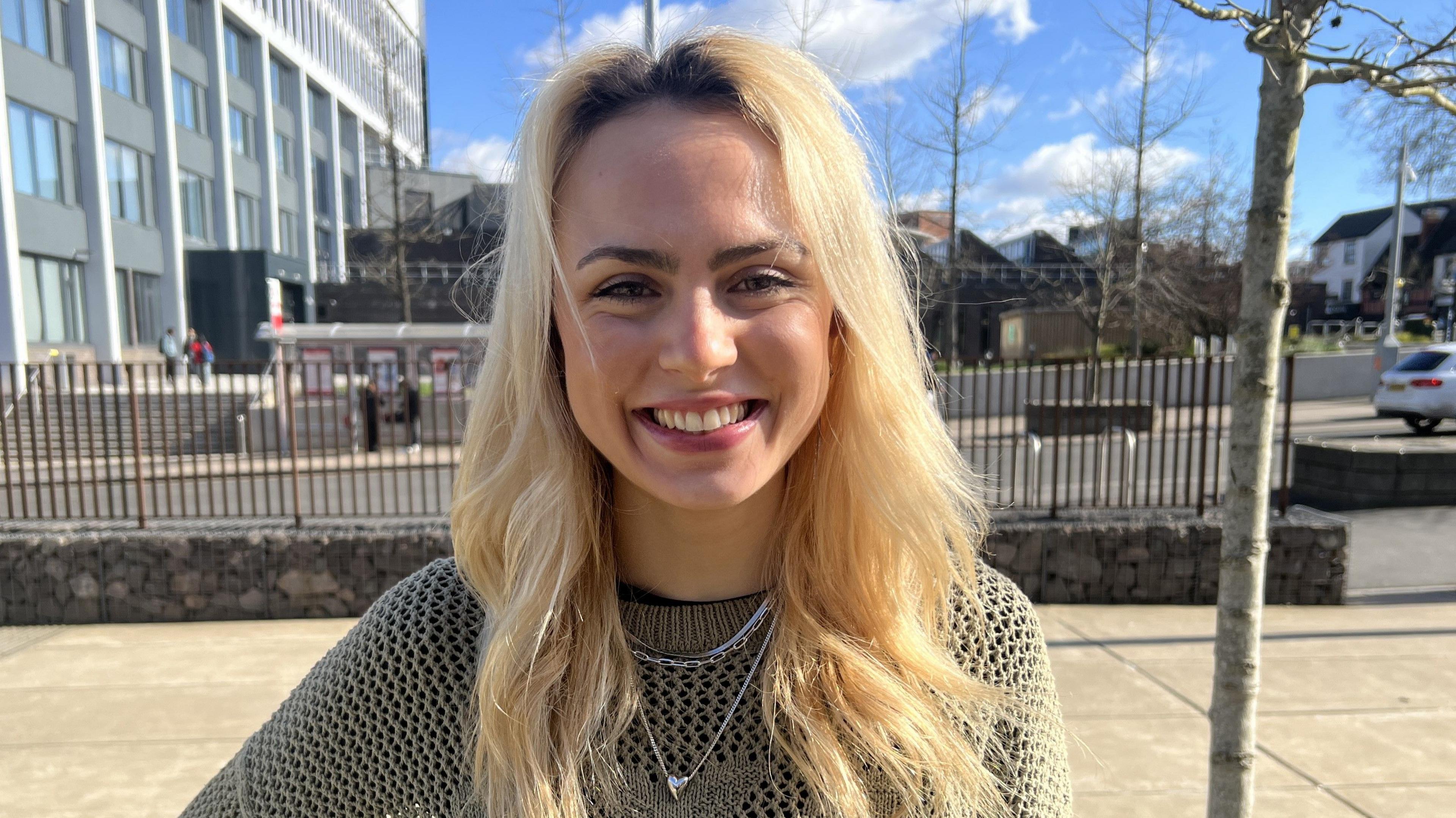 Young woman with dyed blonde hair smiles at the camera in a green netted jumper. She wears what looks like three necklaces, one with a heart on. She is standing on what appears to be a university campus with grey and brown buildings in the background, behind some railings. In the background, a parked car is visible to the right and blurred figures stand on the top of steps to the left. The sky is blue with white clouds in.