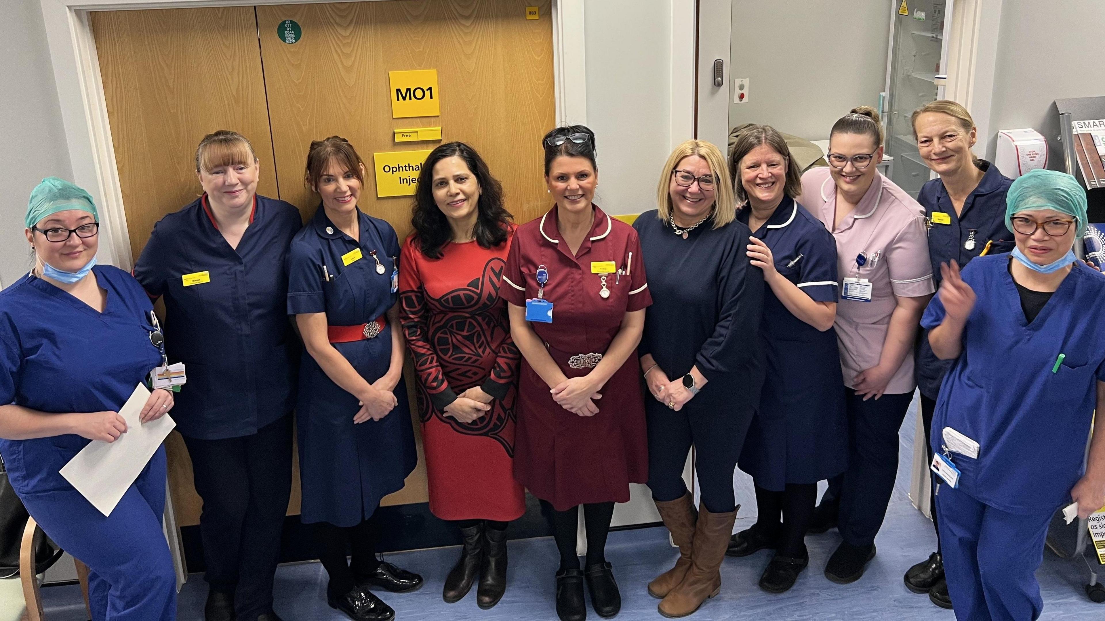 A group of doctors and nurses from the ophthalmology stand together and pose for a photo inside a clinical setting. There are ten people, some dressed in scrubs, others in nursing uniforms and one in a red dress. 