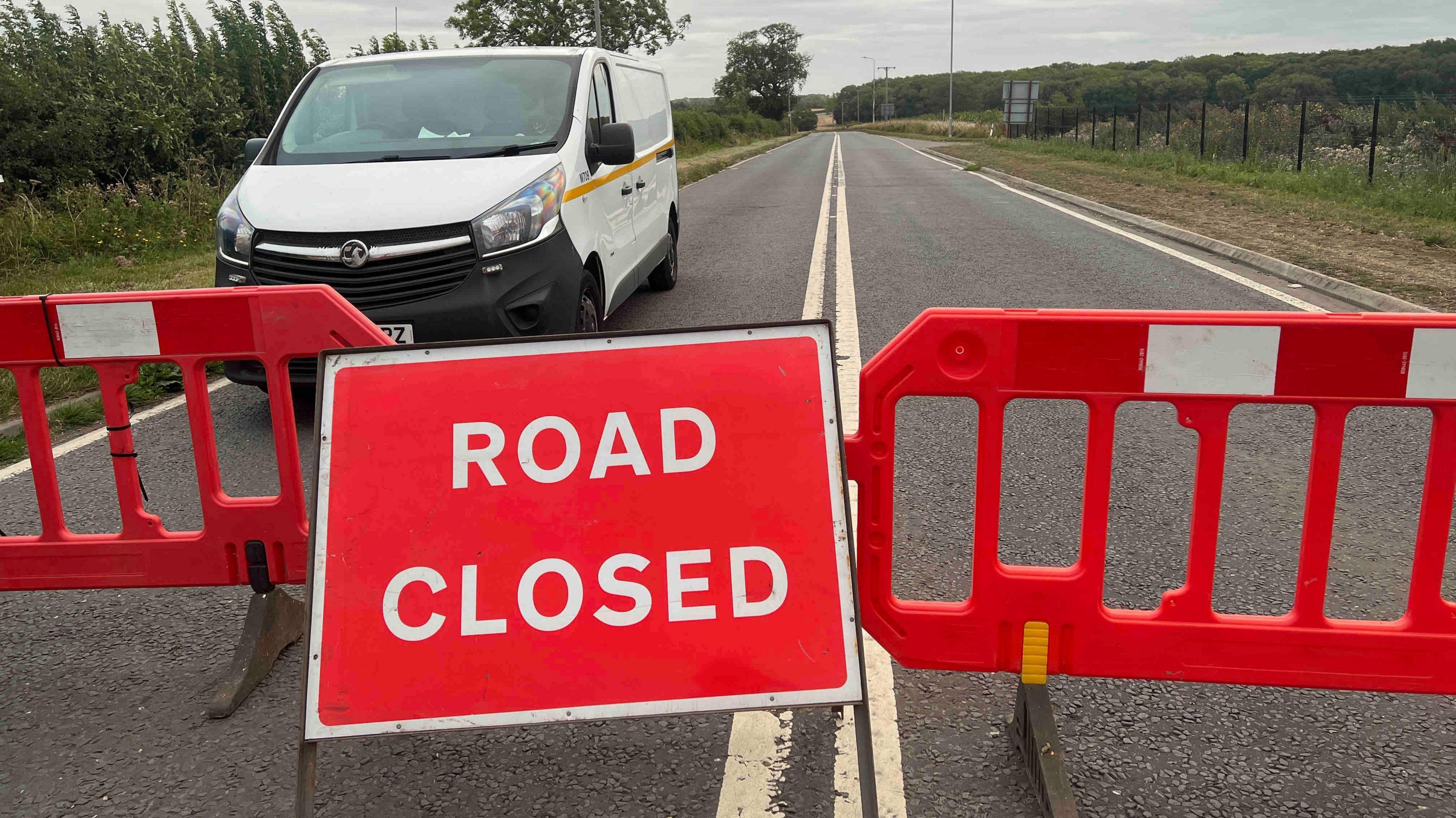 A red 'ROAD CLOSED' sign and red barriers blocking a road; a white van is parked behind the barriers.