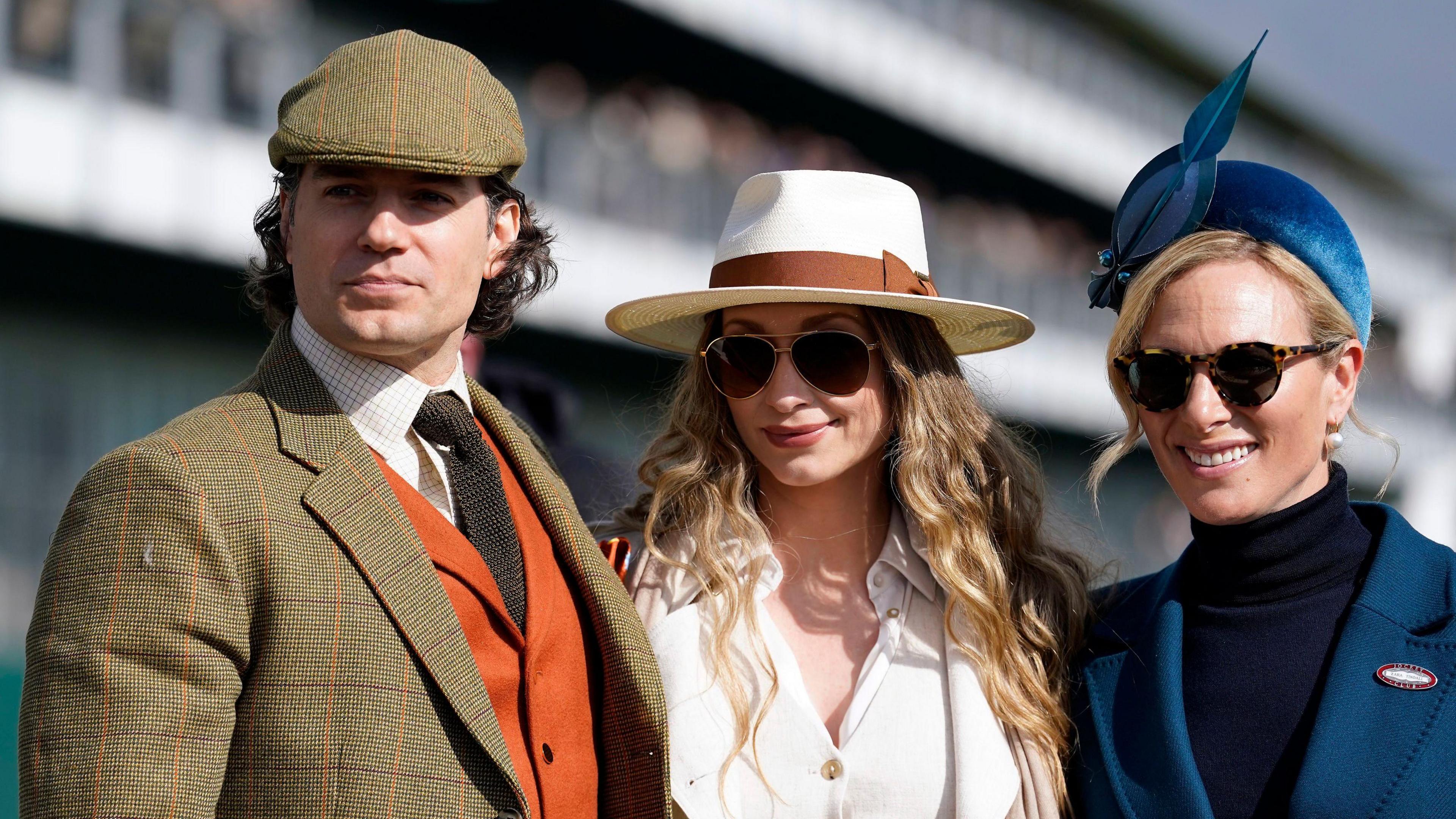 Henry Cavill is on the left and he is wearing a tailored tweed jacket, with a orange waistcoat, white shirt and wool tie. In the middle is Natalie Viscuso, who has long blonde hair, a white sunhat and is wearing a white shirt. Zara Tindall is on the right and is wearing a dark blue turtle neck with a blue jacket on top and a blue fascinator.