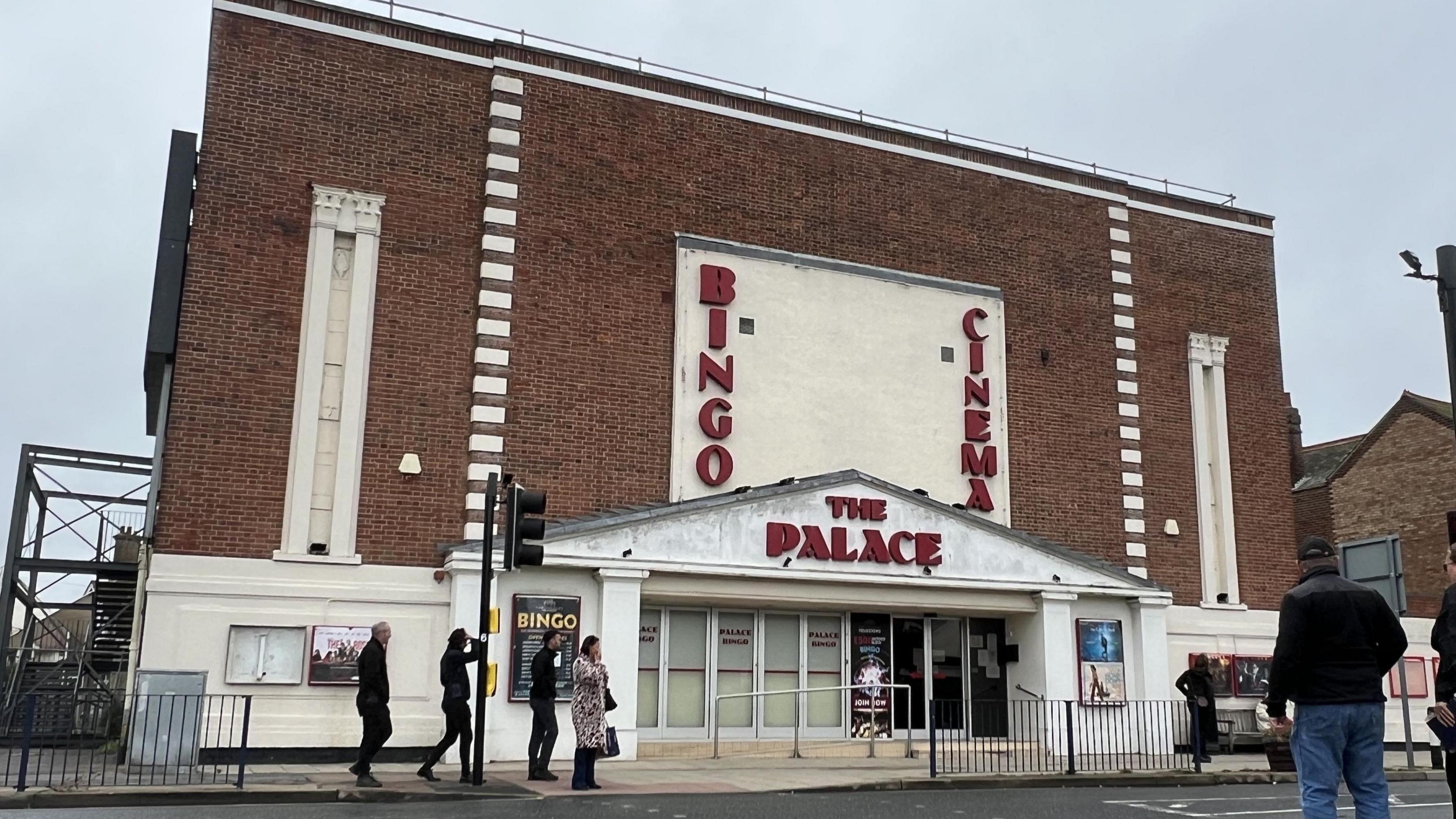A general view of a red brick art-deco building that houses the Palace Cinema. Residents walk on the pavement outside of it.