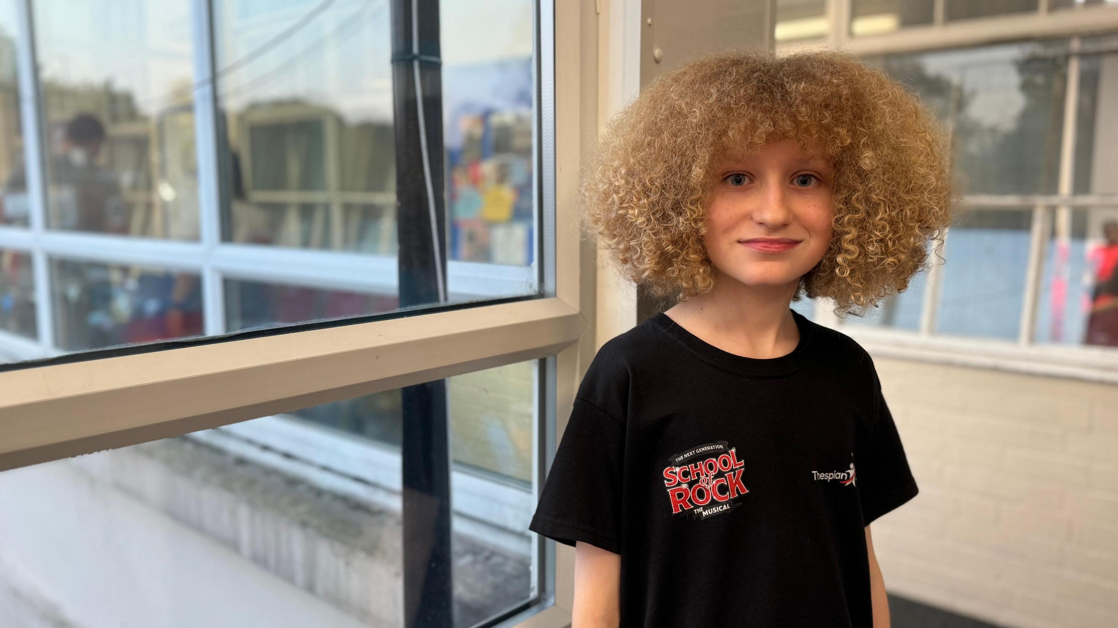 Ben, standing in a glassed corridor at the centre in Brownhills, looks at the camera. He is wearing a black School of Rock T-shirt