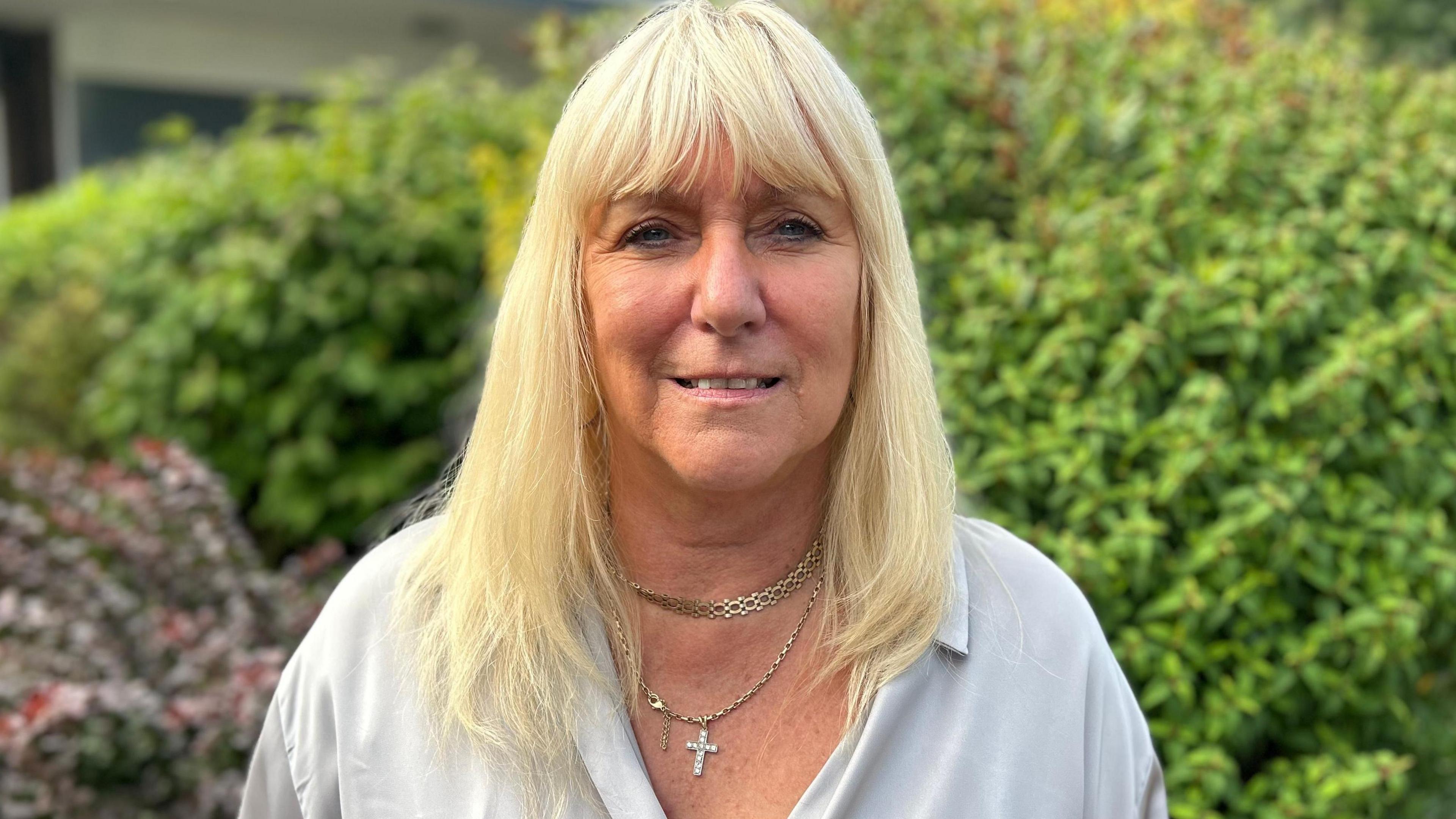 Julie Stokes, who has blonde hair and is wearing gold necklaces and a silver shirt, standing in front of green bushes.