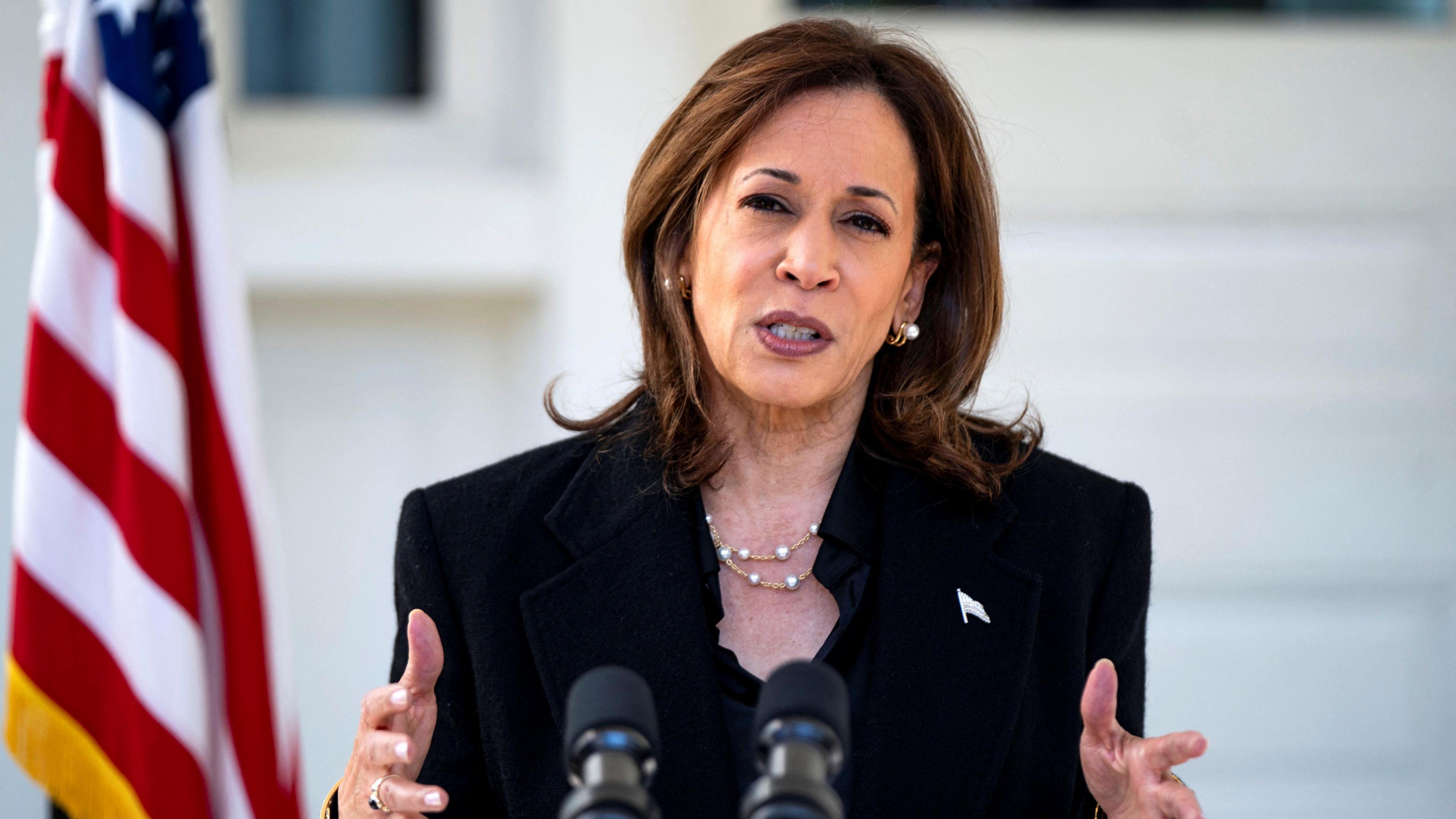 Kamala Harris stand in a black suit with her hands forward with the US Naval Observatory in Washington, U.S. in the background and an American flag 