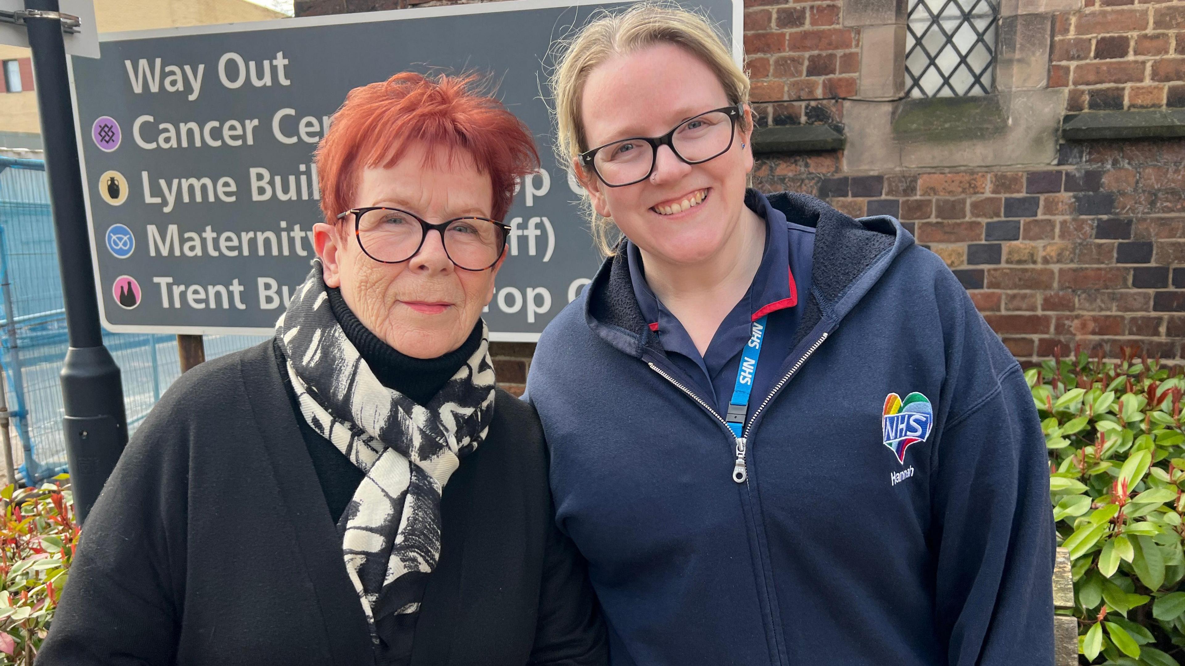 A woman with short red hair and glasses stands next to a woman with blonde hair and glasses. They are both in front of a sign and a bush outside the entrance to a hospital building.