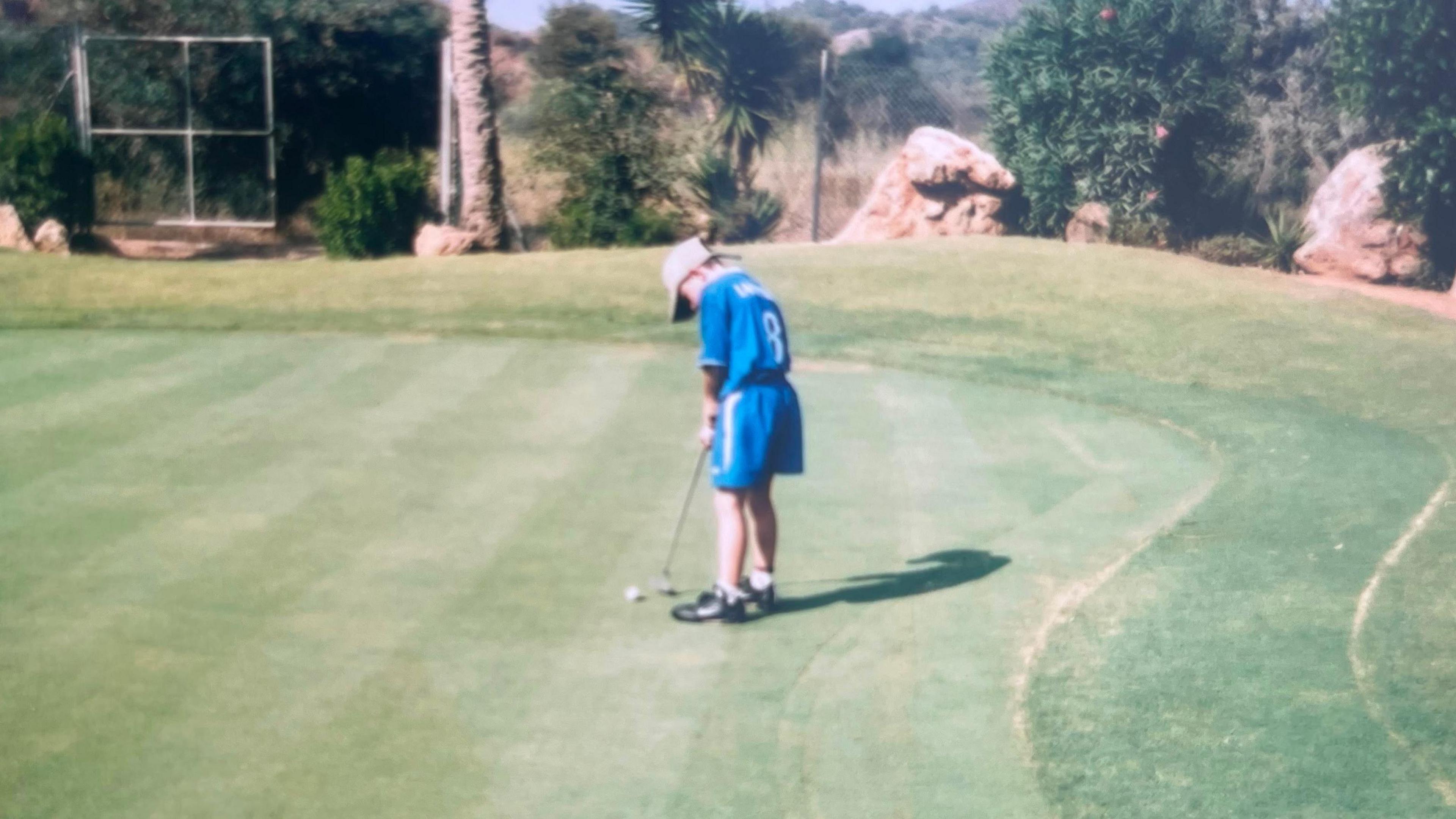 Tom Sloman, as a child and dressed in a blue football kit with a grey baseball cap, putting the ball on the green.