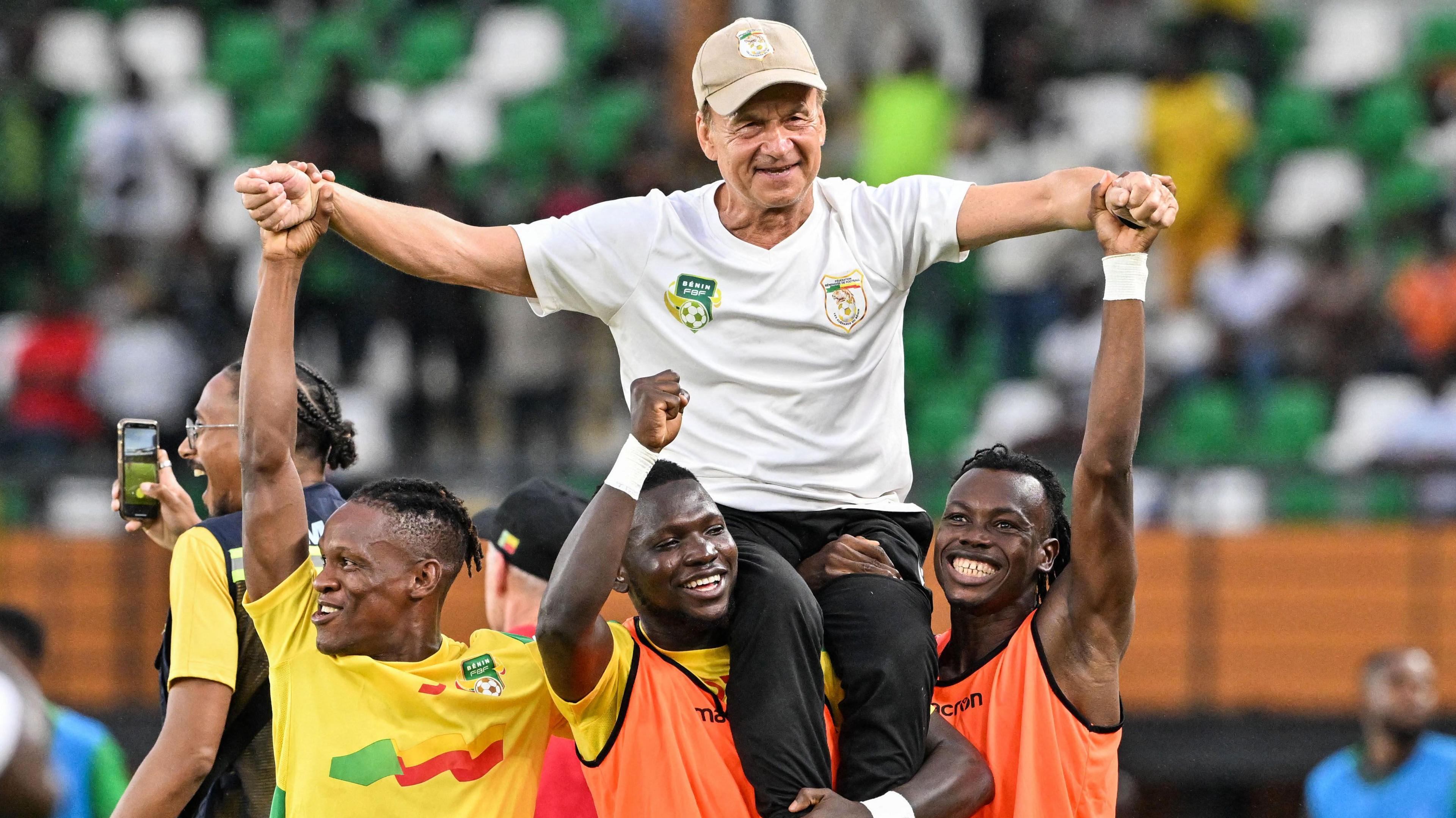 Gernot Rohr wearing beige cap, black trousers and white Benin training top is raised on the shoulders three smiling Benin players as they all raise their arms in celebration after beating Nigeria