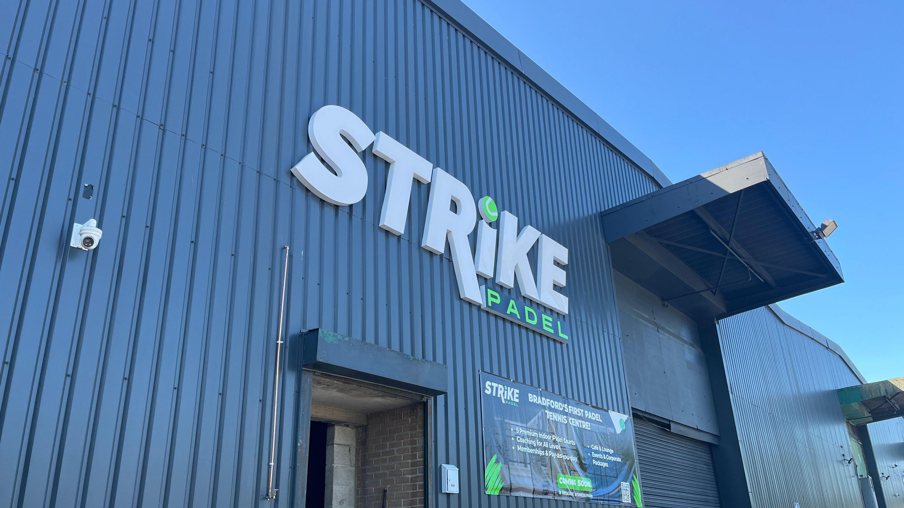 Exterior of Strike Padel - a blue warehouse-type building with signage advertising the company on the side.
