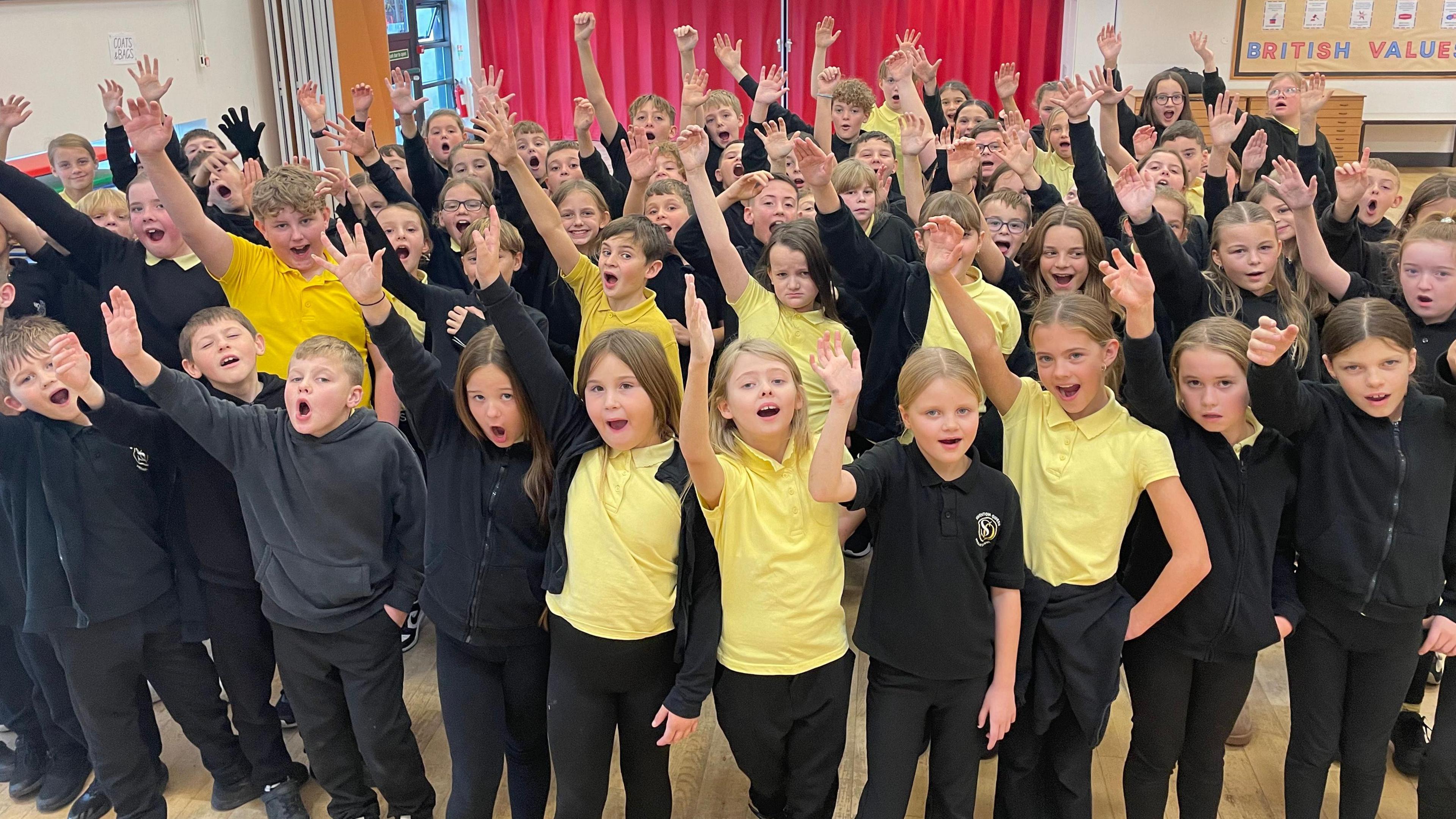 A large group of schoolchildren in black uniforms with yellow polo shirts all have one hand in the air and are looking towards the camera. Their faces show they are singing. They are indoors in a hall with red curtains in the background and a noticeboard that says British Values on it.
