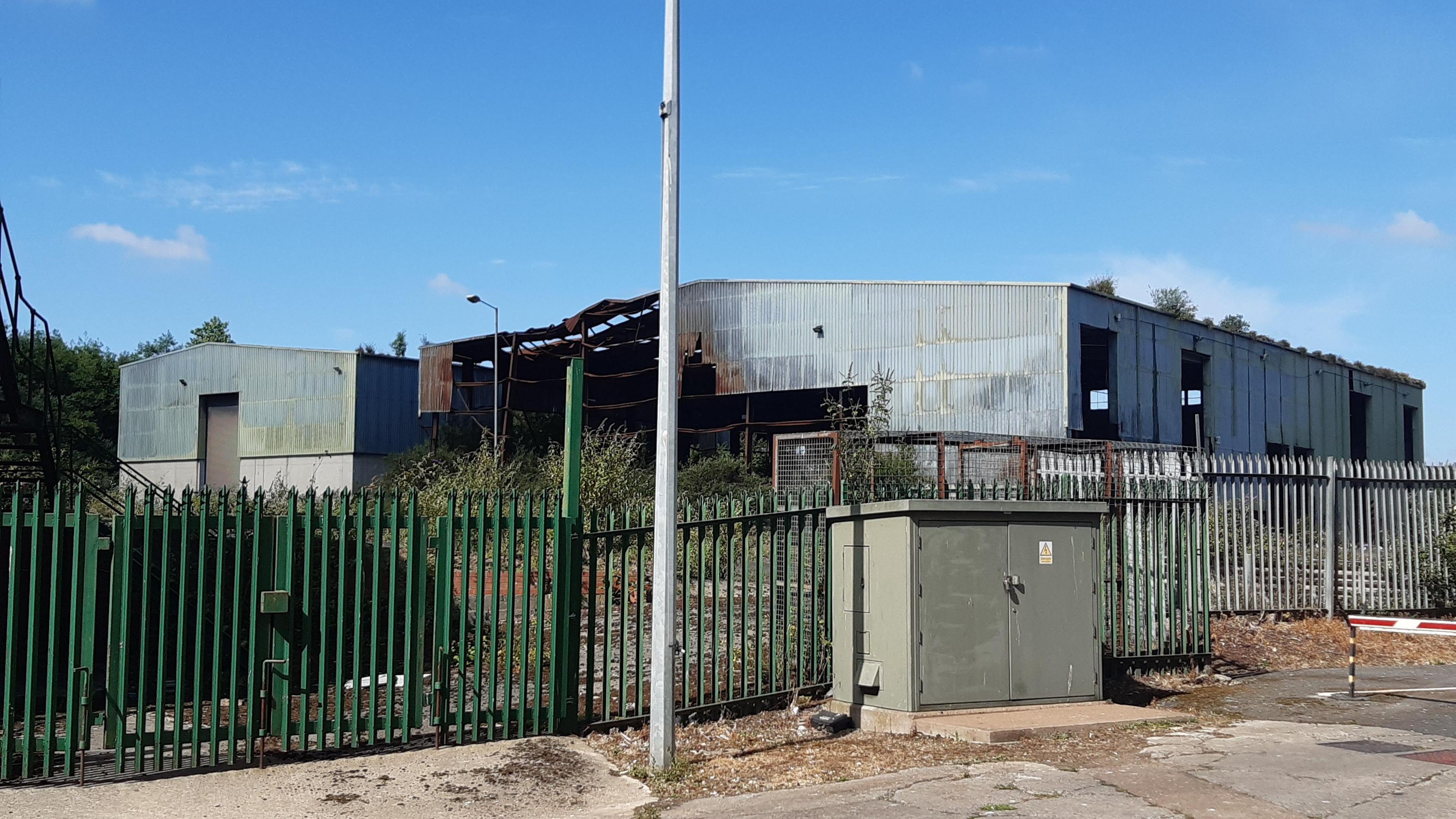 The scorched grey panel buildings of the former Alex Smiles Ltd  waste management site, photographed in July 2025. The structures are behind a green metal fence.