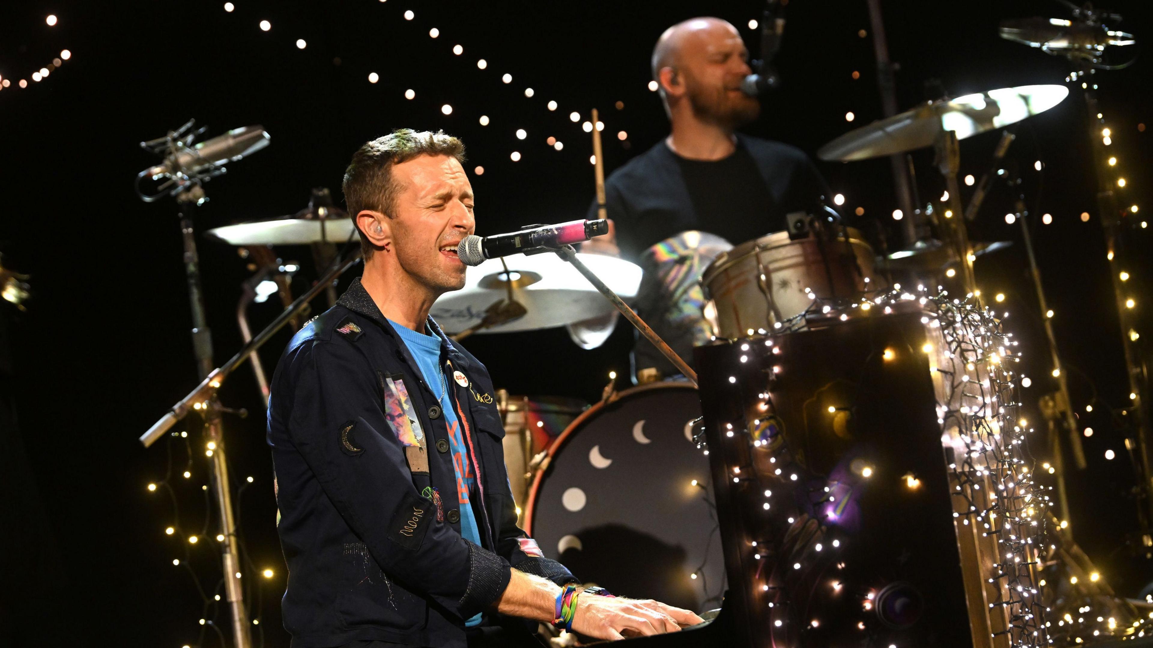 Chris Martin sits at and plays a piano while  singing into a microphone. He's wearing a navy blue jacket with blue T-shirt. Behind him is a drummer and drum kit. The stage is covered in fairy lights