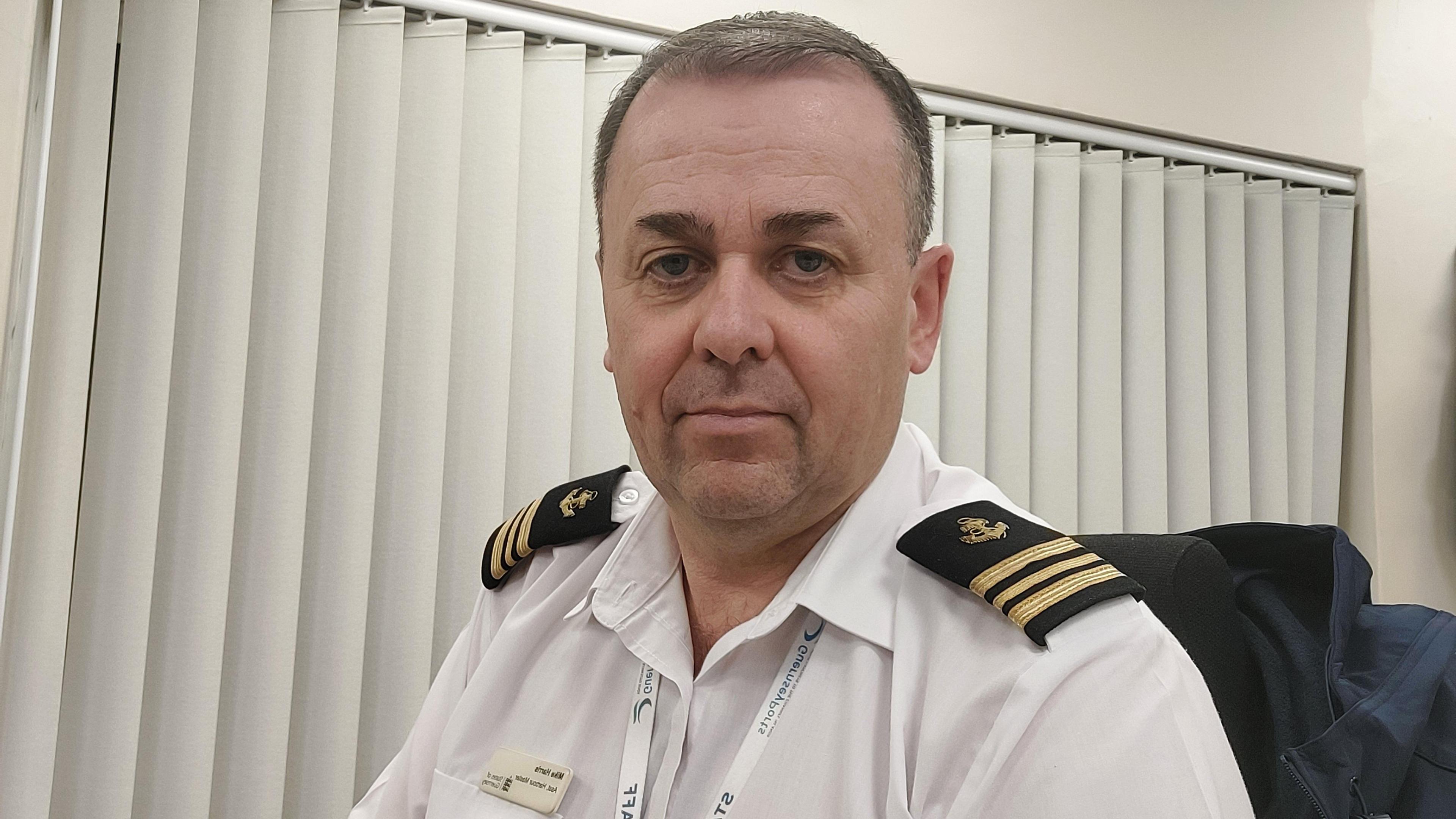 Guernsey Ports assistant harbour master Mike Harris sits in an office on a chair with a blue coat hung on the back of it. He is wearing a white shirt with black epilates with three gold stripes on them. A window with white blinds drawn across it is in the background. 