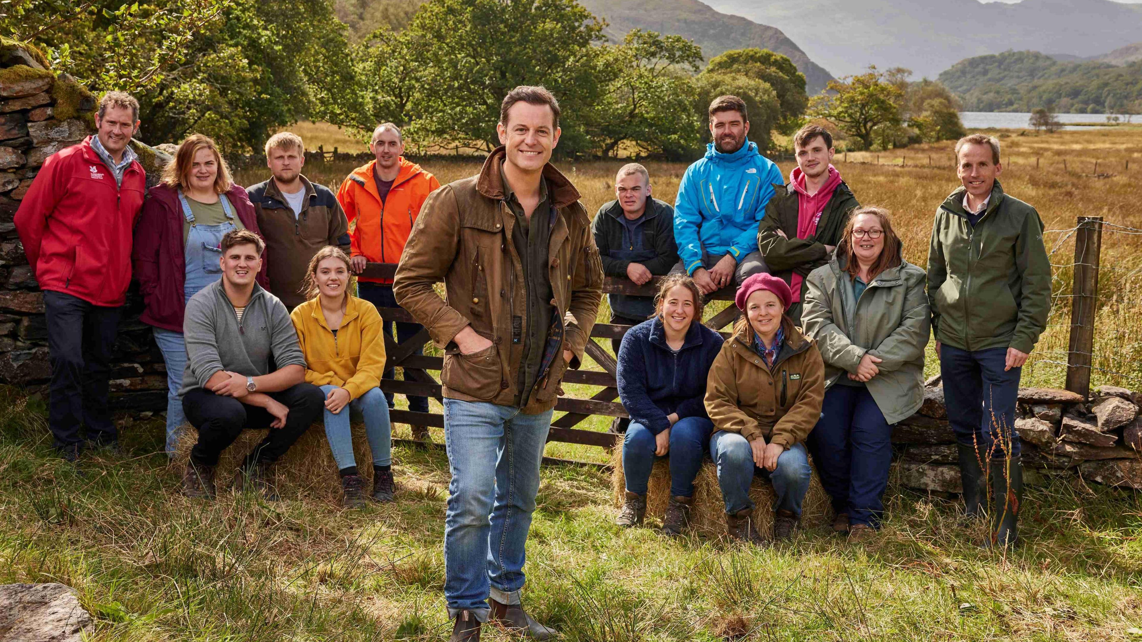 Matt Baker stands in front of the competitors and judges in Channel 4's Our Dream Farm. He is wearing a brown coat and blue jeans. The competitors and judges are either stood or sat behind him and have jeans on and different coloured coats. There are both male and female participants.