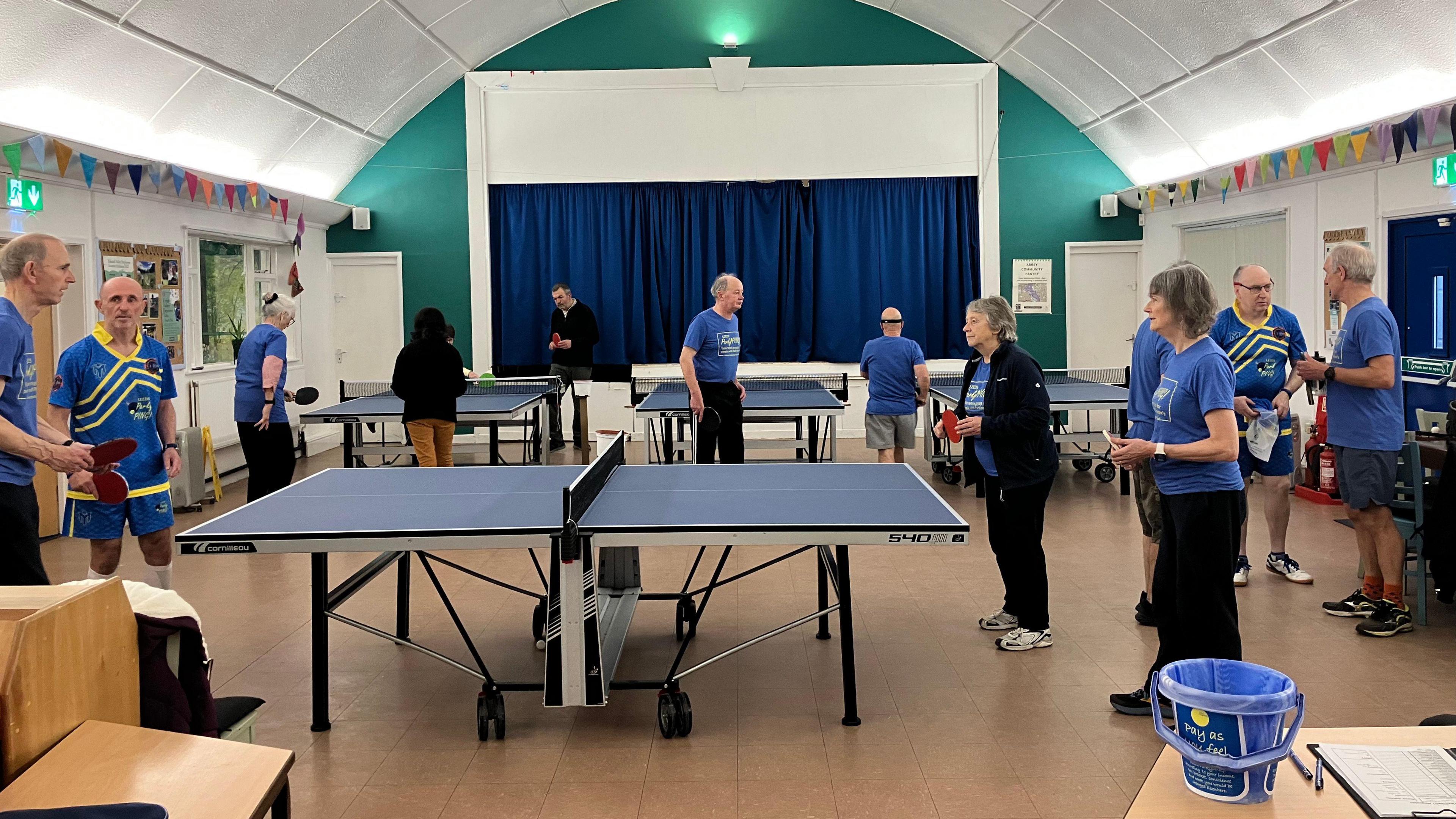 A large indoor hall with a high arched ceiling and bright lighting, set up for table tennis activities. Several tables are arranged in rows, and multiple people wearing blue sports shirts are standing around or playing. 