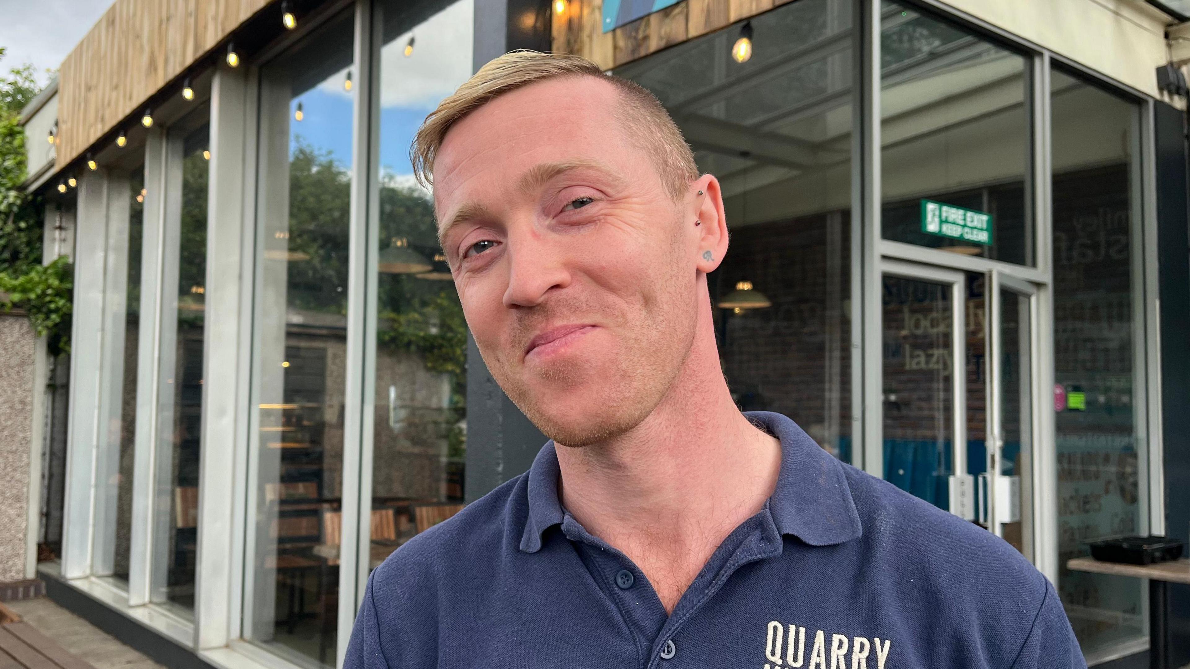 Richard pictured looking into the camera and smiling slightly. He's wearing a navy blue polo shirt, on the left hand side it has 'QUARRY' embroidered in beige. He's stood in front of the entrance to the Quarry Kitchen, which has large floor-to-ceiling windows and fairy lights strung along the roof edge.