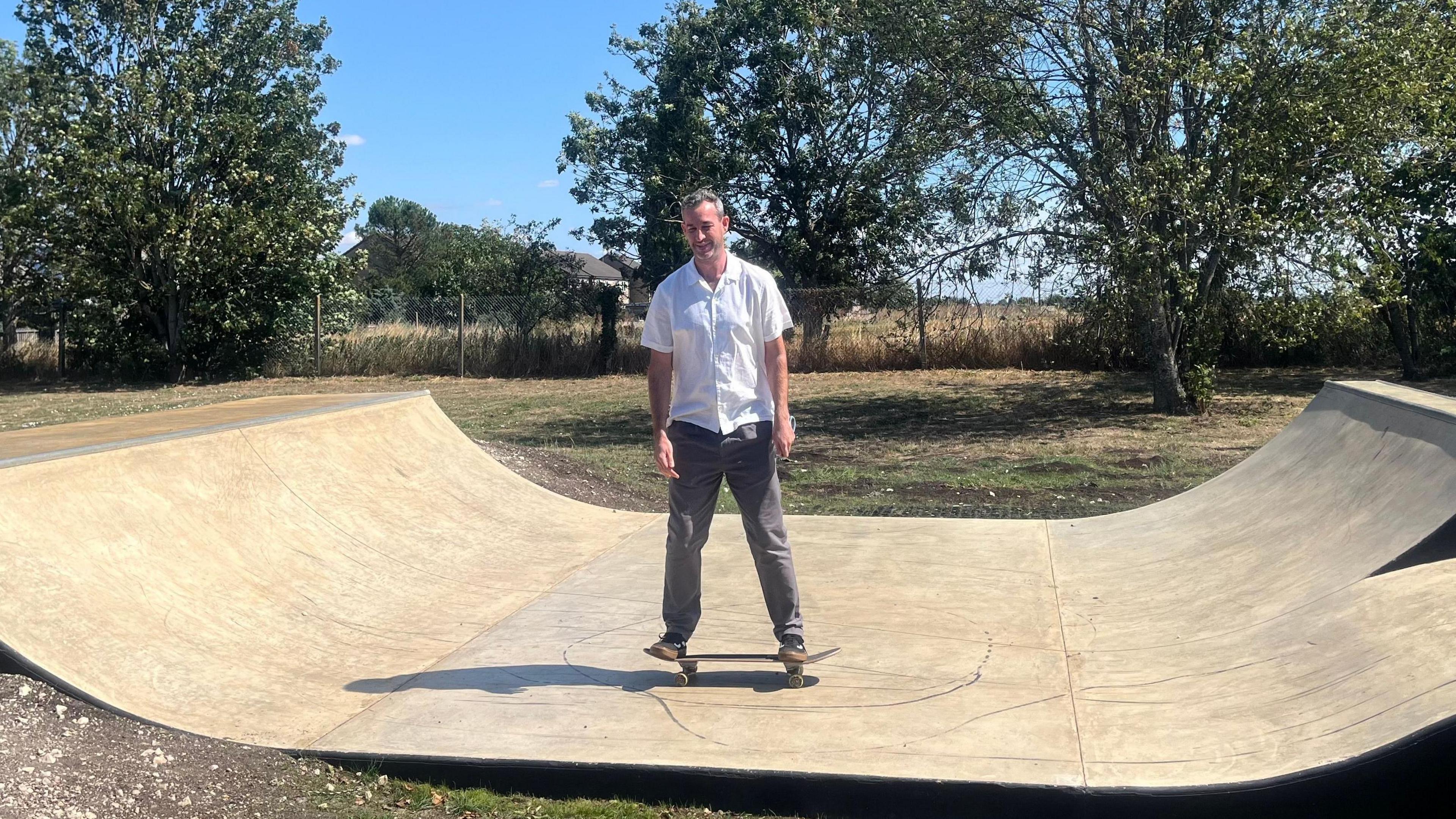 Mark Ormston is trying out the new skate park. He is standing in the dip of the ramp on top of a skateboard with his arms down by his sides. He is smiling while wearing grey jeans and a white shirt. It is a sunny day with blue skies.
