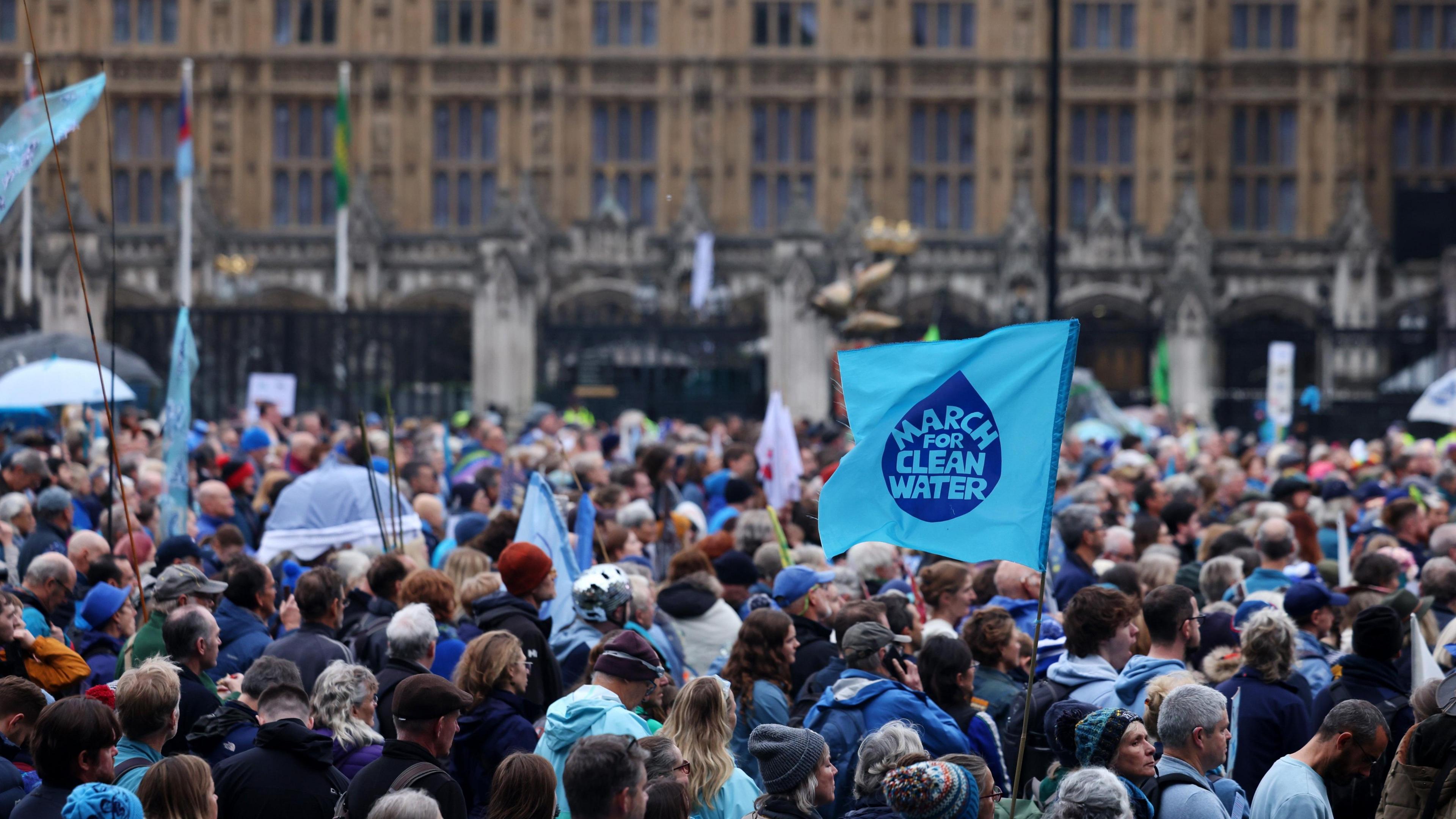 A crowd of protesters outside the Houses of Parliament. A blue flag is held up, with the words March for Clean Water written on it.