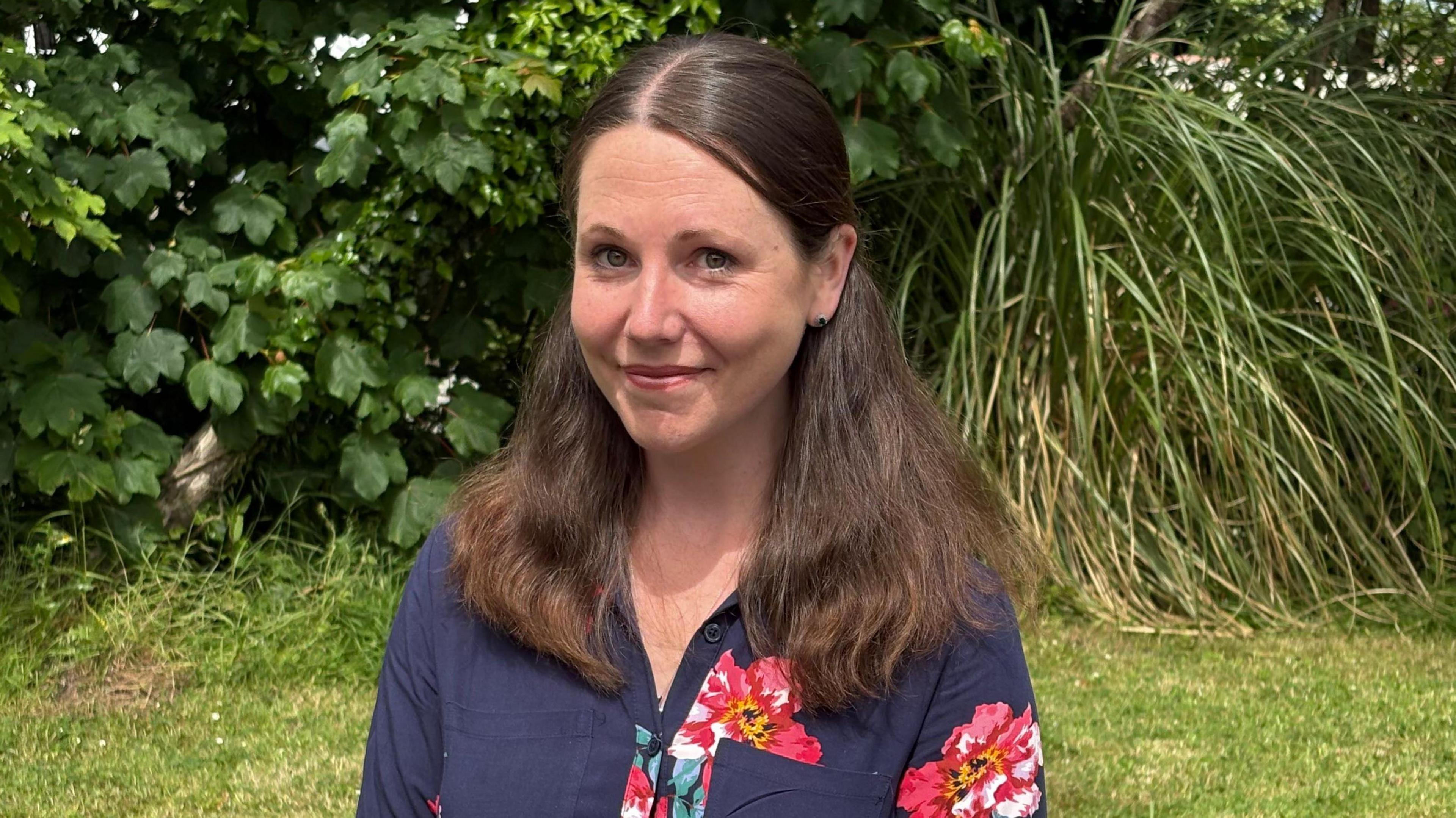 A young woman with long dark hair over both her shoulders wearing a dark blue top with flowers on it, standing in front of a hedge in a garden.